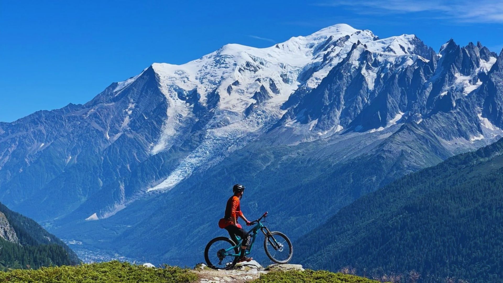 A man on a mountain bike stands on a rock, gazing at the expansive mountain range in the background.