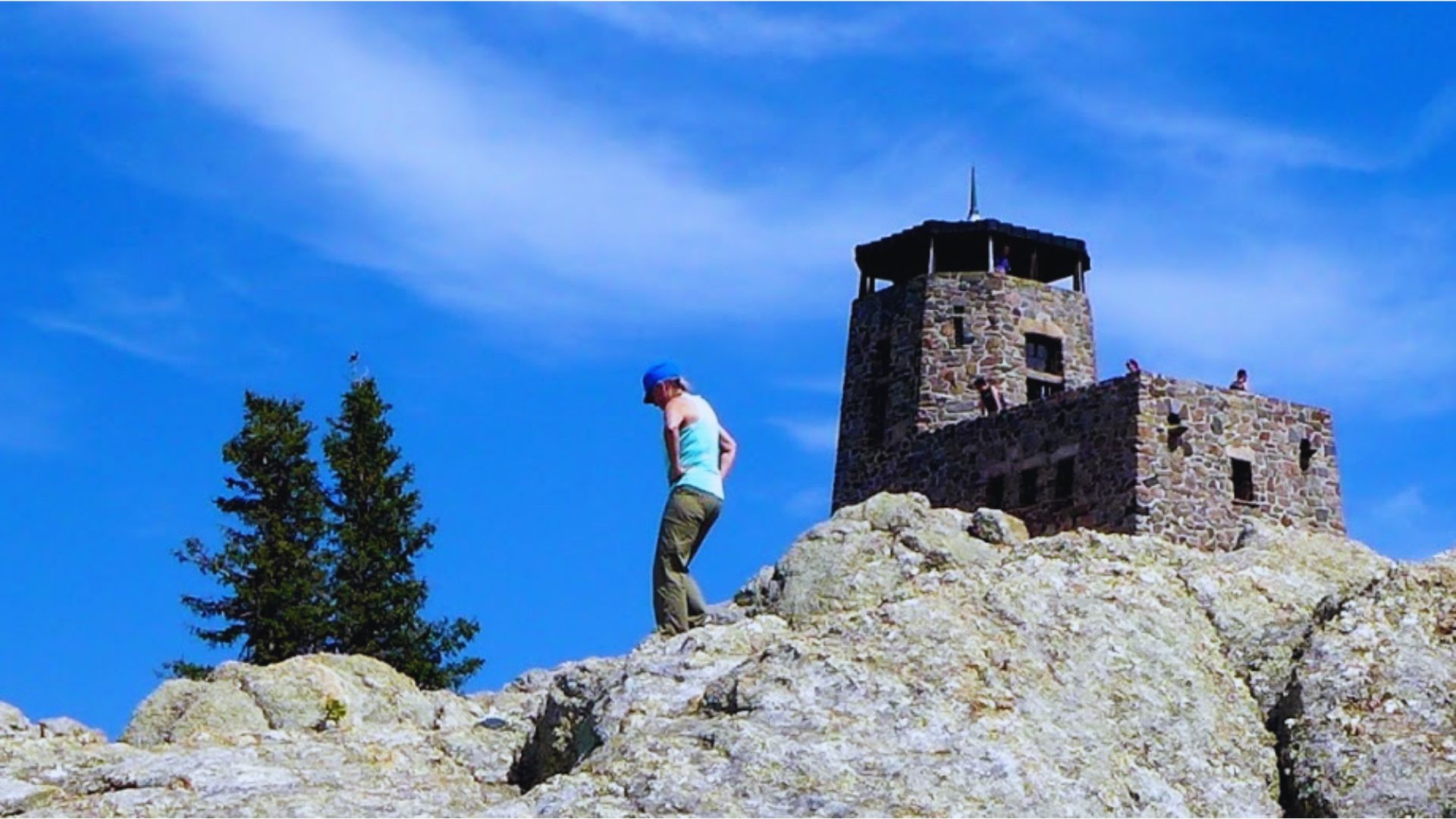 A person poses on a rock with a distant tower rising in the background under a bright sky.