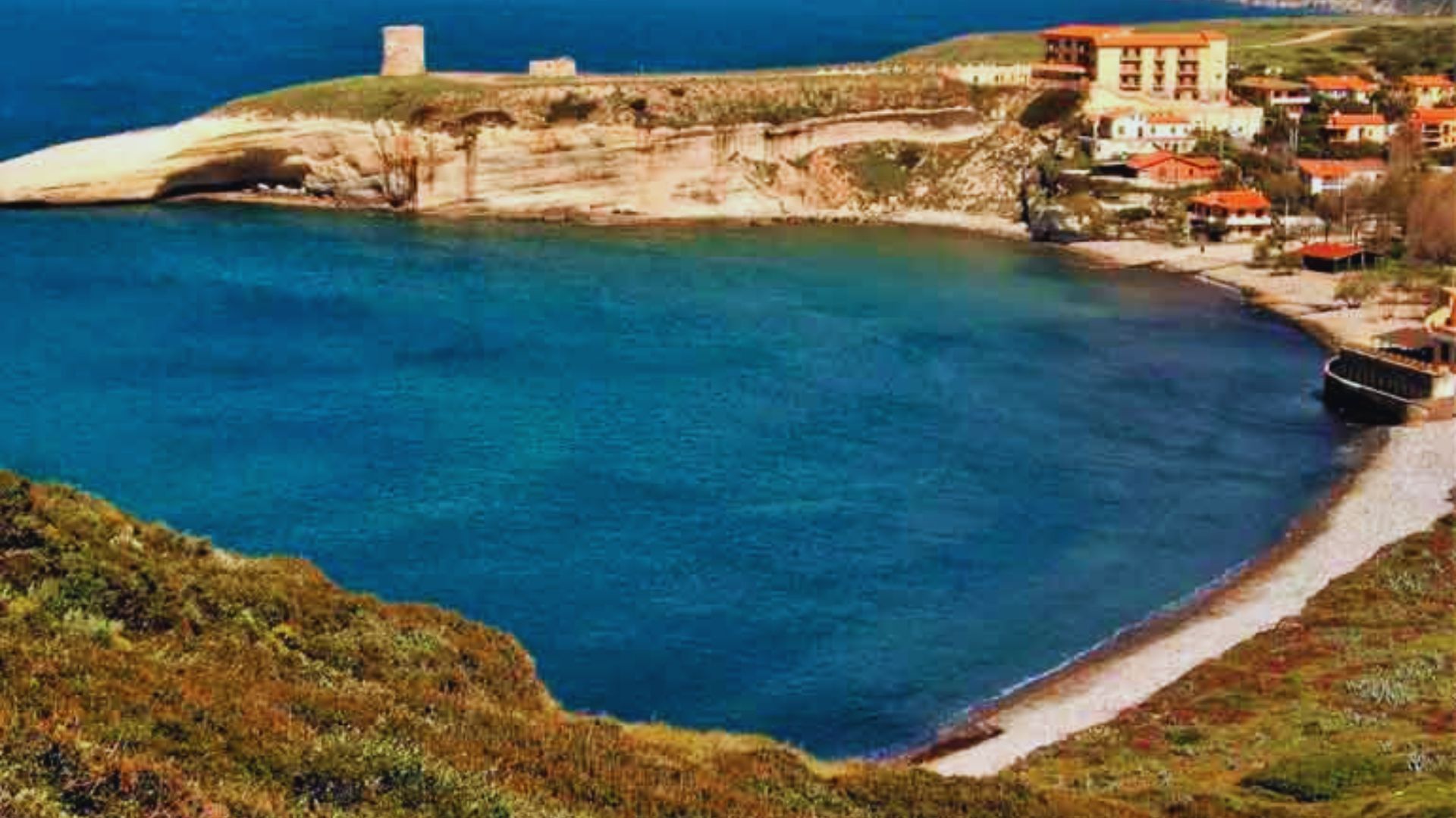 A serene coastal landscape features a crescent-shaped turquoise bay bordered by lush green cliffs. Red-roofed buildings sit on a hill, evoking a peaceful vibe.