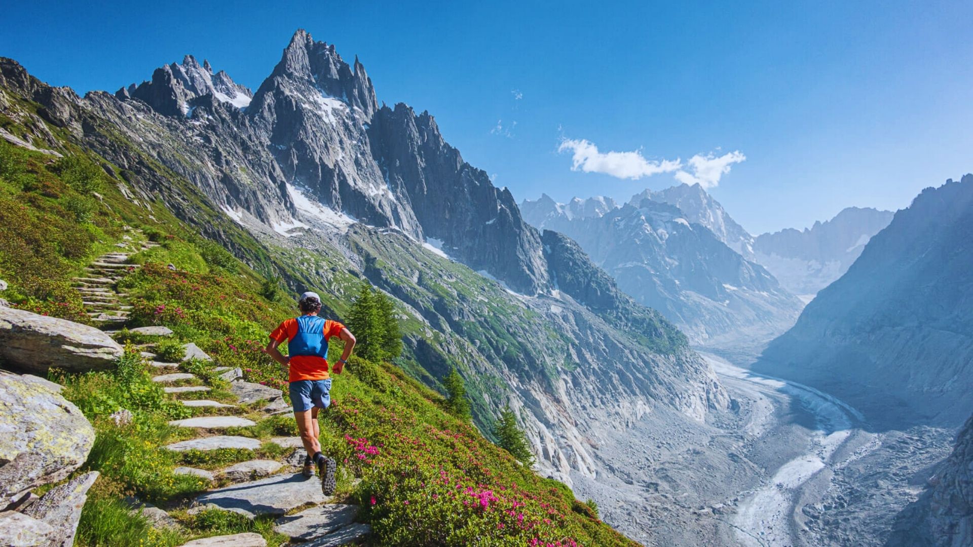 A man trekking along a mountain path, with steep slopes and vibrant greenery flanking the trail under bright sunlight.