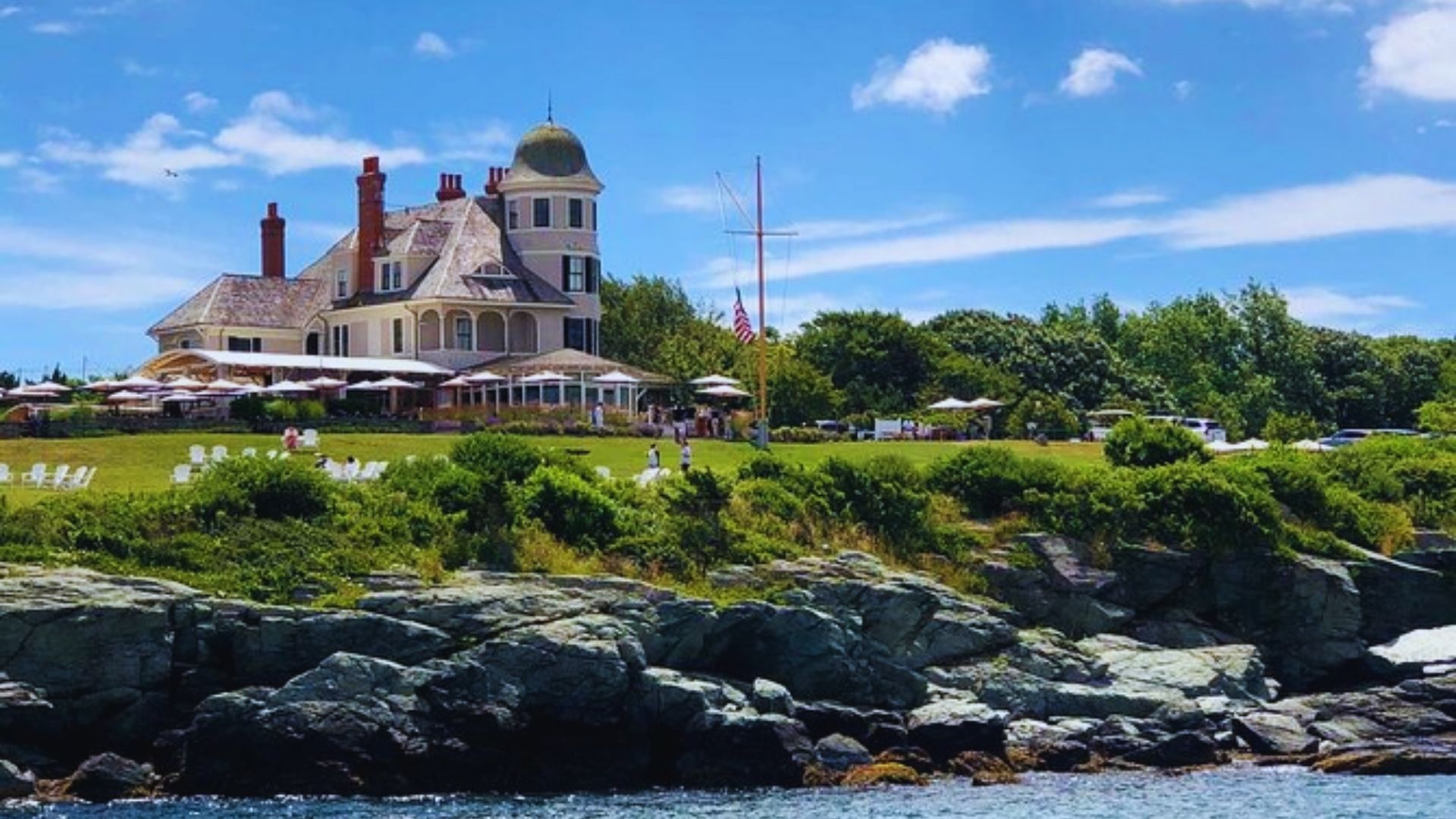 A large house stands on the shore, overlooking a tranquil body of water under a clear blue sky.