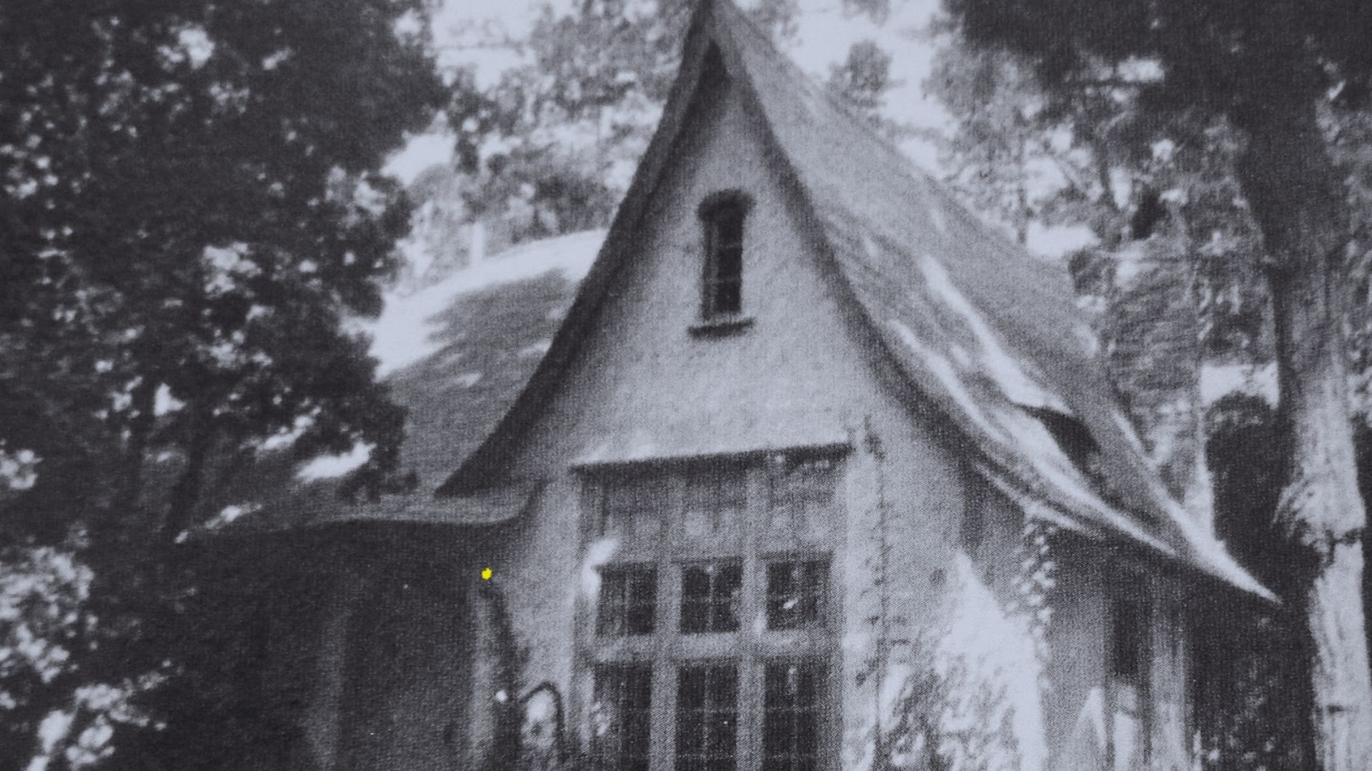 Black and white photo of a quaint, storybook-style cottage with a steep, pointed roof. Surrounded by tall trees, the scene feels nostalgic and serene.