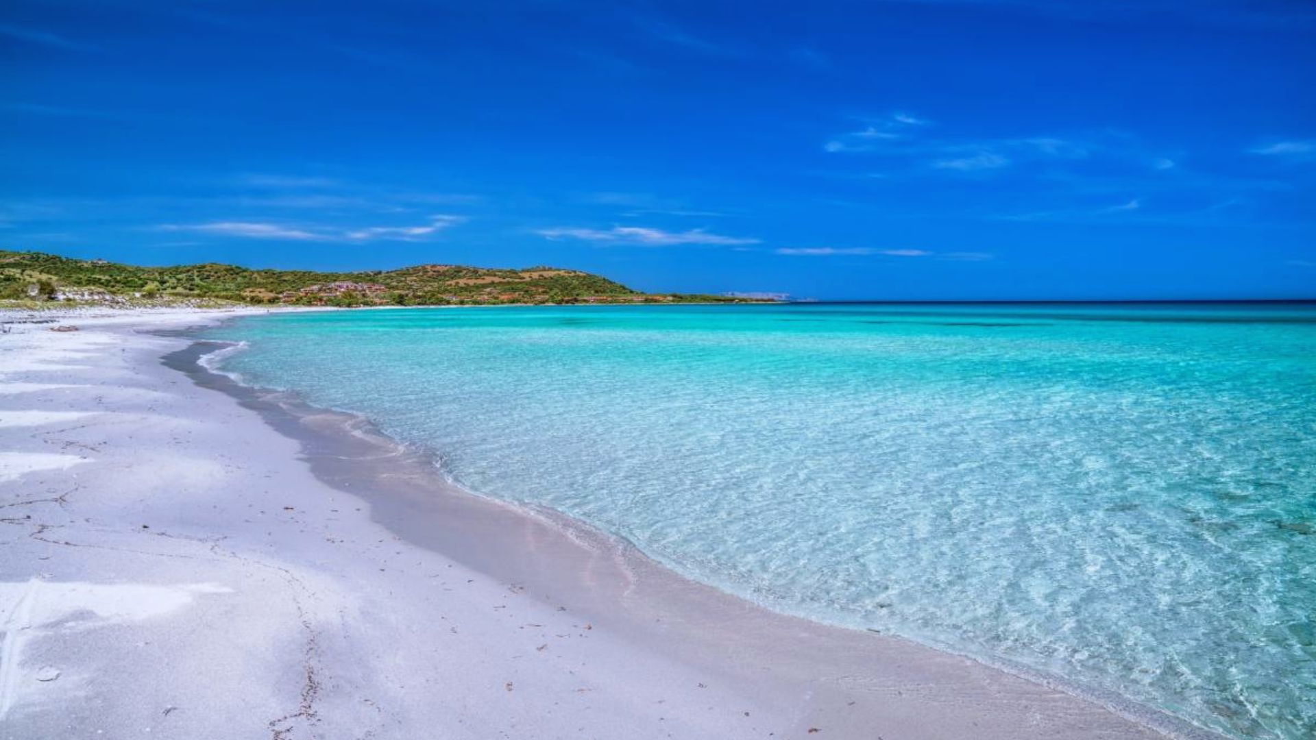 A serene beach with white sand curves gently along vivid turquoise water under a clear blue sky. Distant green hills border the horizon, evoking tranquility.