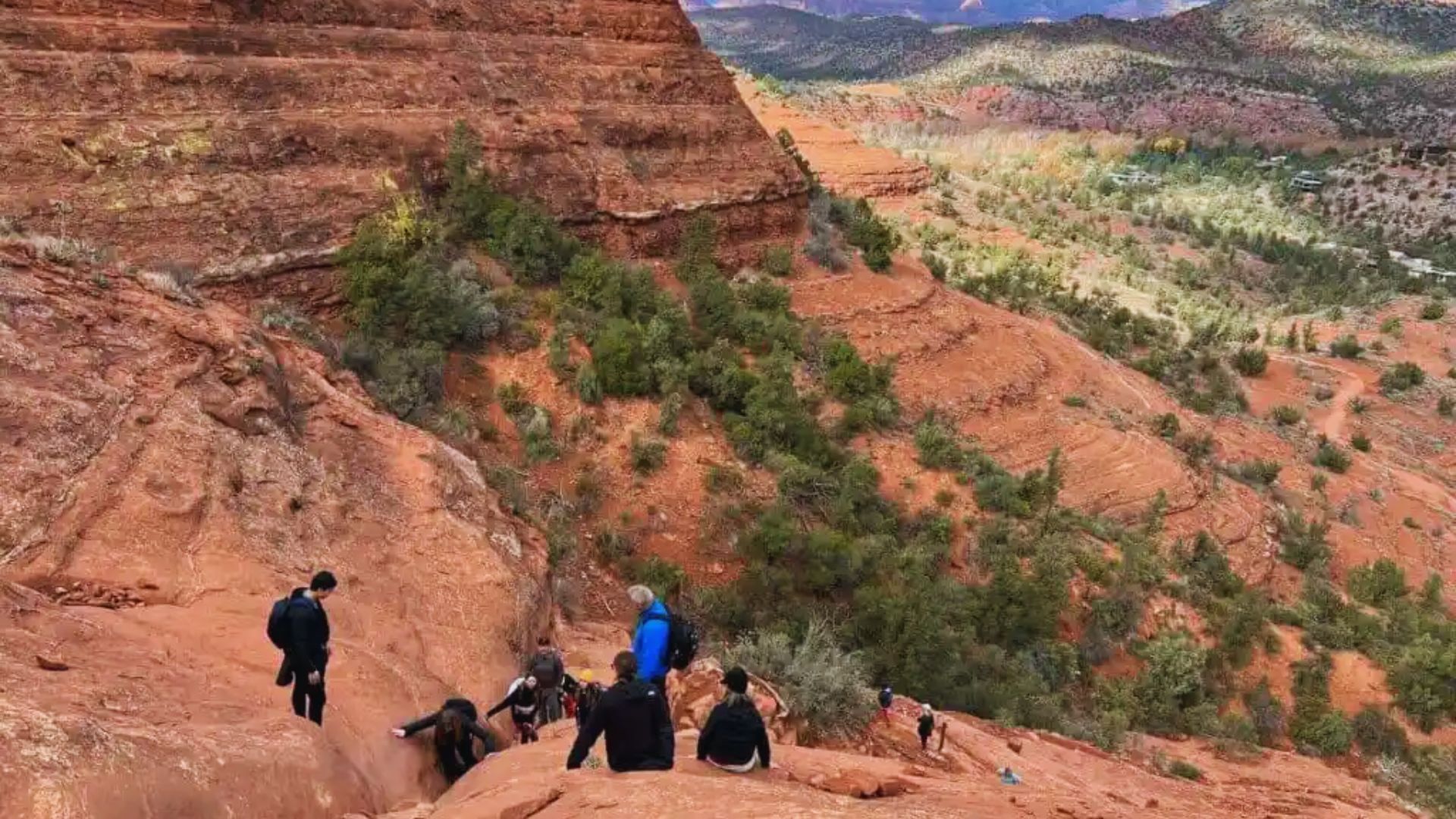 A group of hikers climbs a steep, rocky red cliff, surrounded by sparse greenery. The expansive, vibrant landscape stretches under a vast sky.