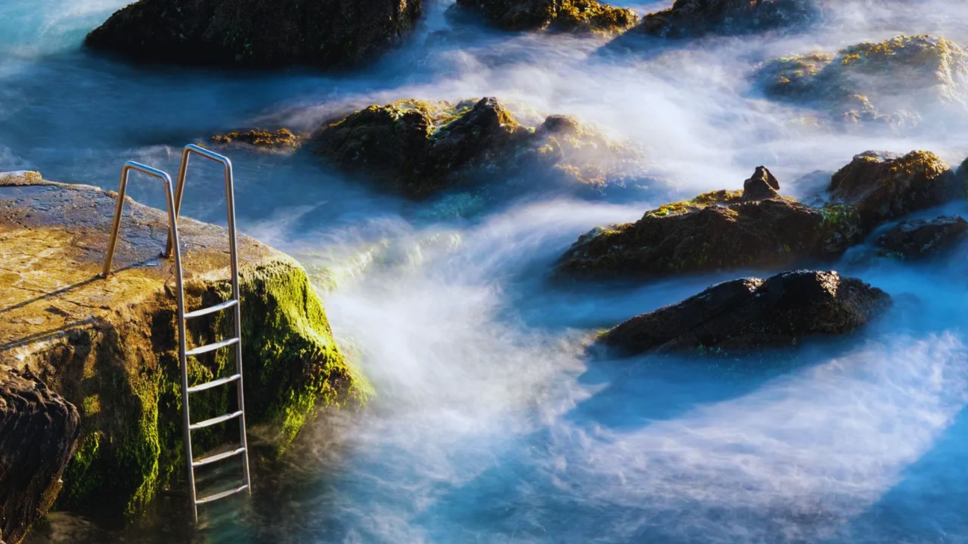 A ladder rests on rocky terrain beside the ocean, with waves gently lapping at the shore in the background.