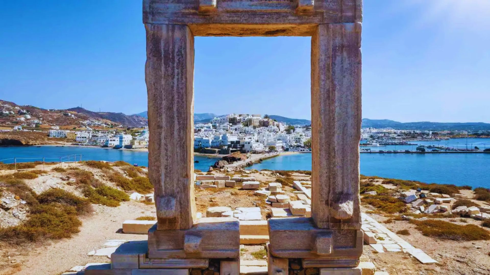 View through an ancient stone archway frames a distant Greek island village against a bright blue sky and sea, conveying a sense of history and serenity.