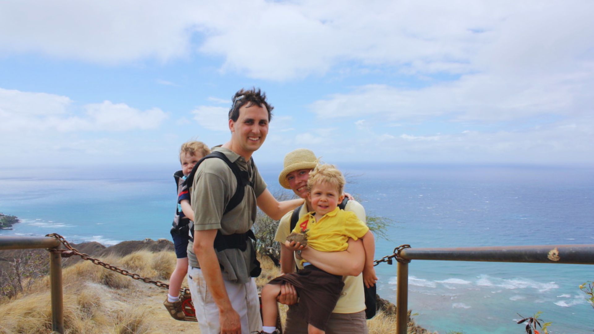 A man and two children stand on a railing, gazing out at the ocean under a clear blue sky.