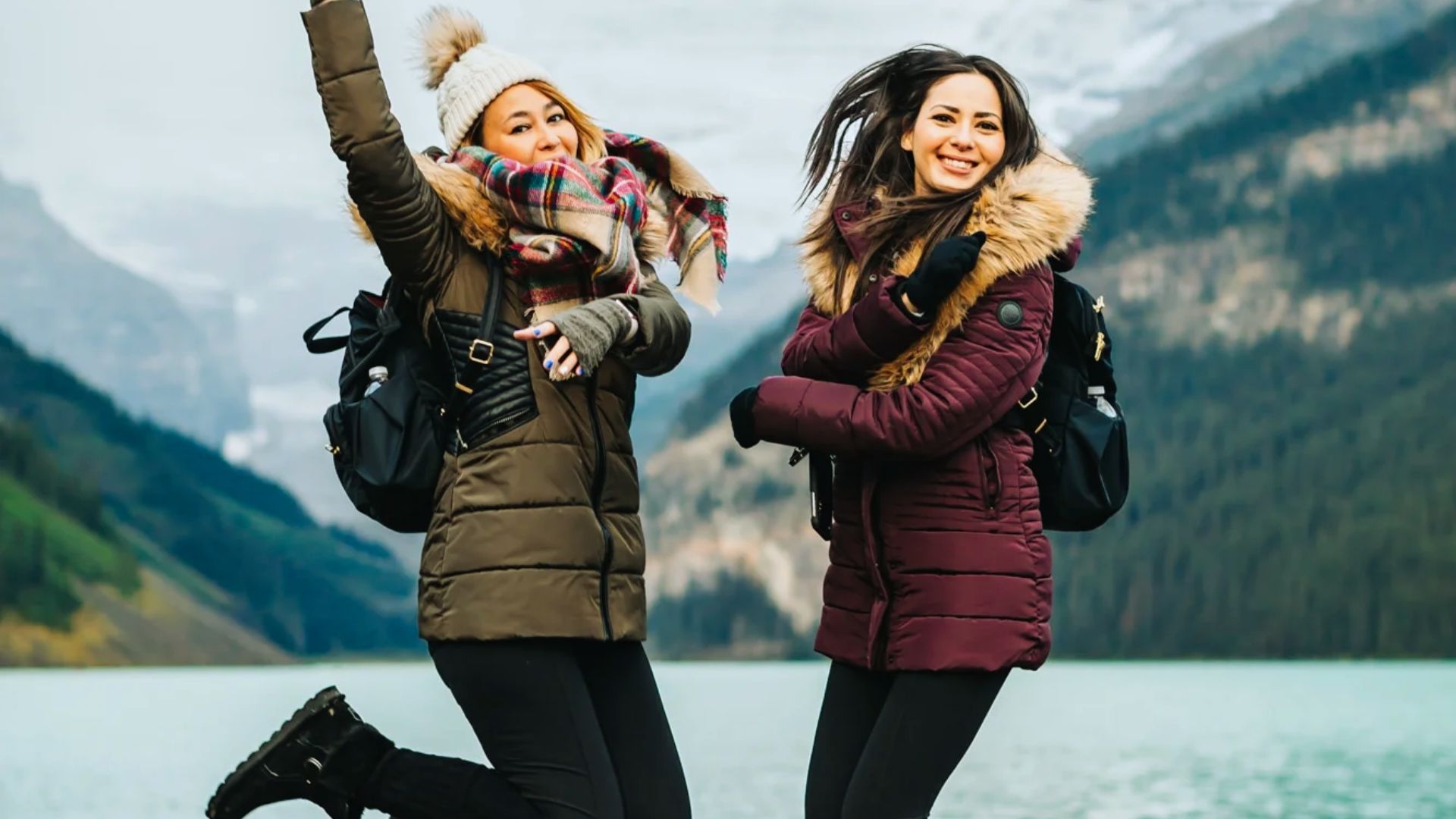 Two women in winter jackets joyfully jump in front of a scenic, misty mountain lake. Both wear backpacks, exuding excitement and adventure.