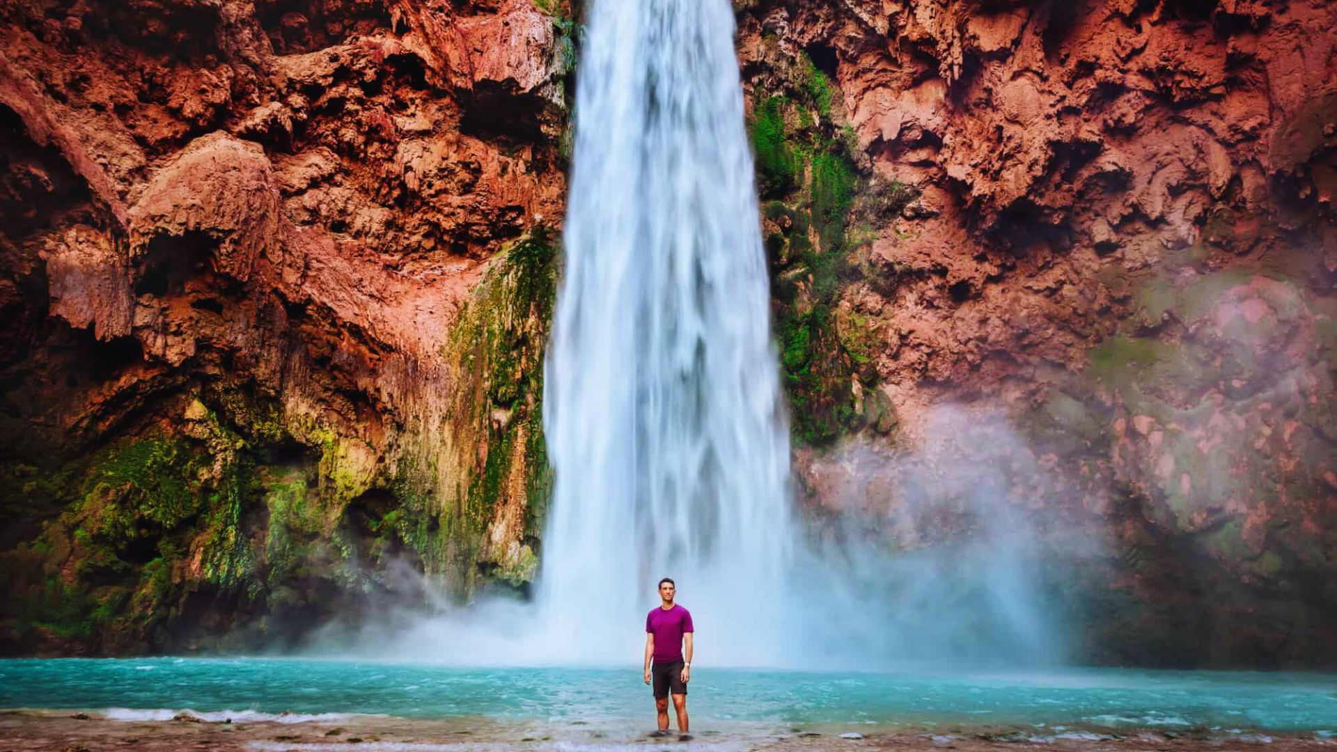 A person in a purple shirt stands in front of a tall waterfall cascading down red rocky cliffs into a turquoise pool, creating a serene, majestic scene.