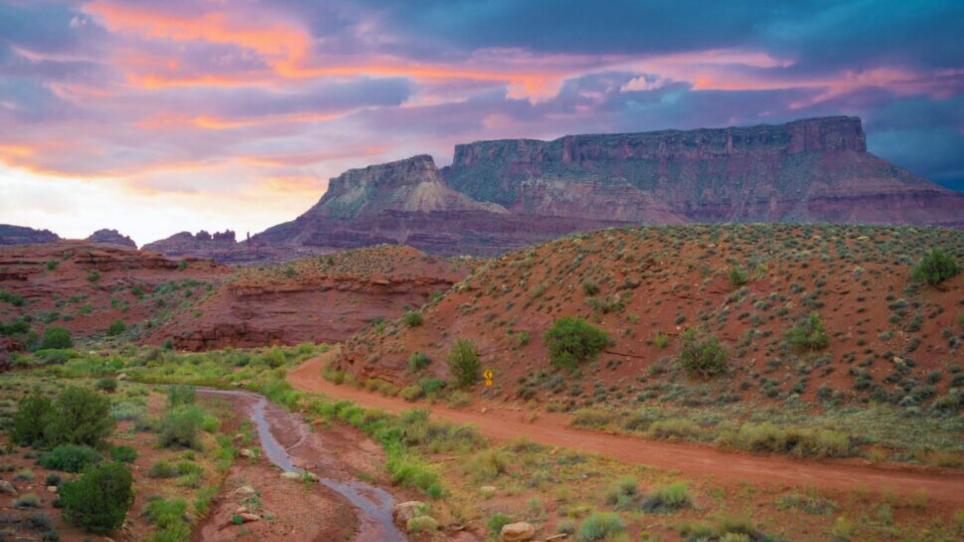 Sunset over a desert landscape with red rock formations, green shrubs, and a winding path. The sky is filled with vibrant orange and purple hues.