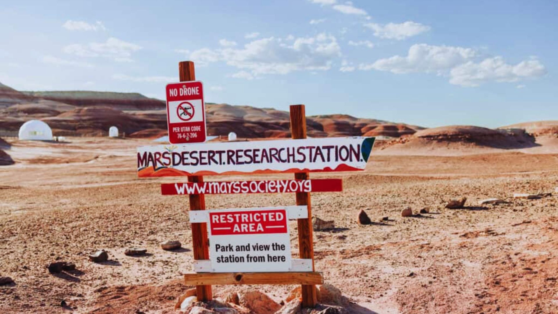 Sign for Mars Desert Research Station in a rocky, arid landscape under a clear blue sky. Dome structures in the background. Restricted area notice.