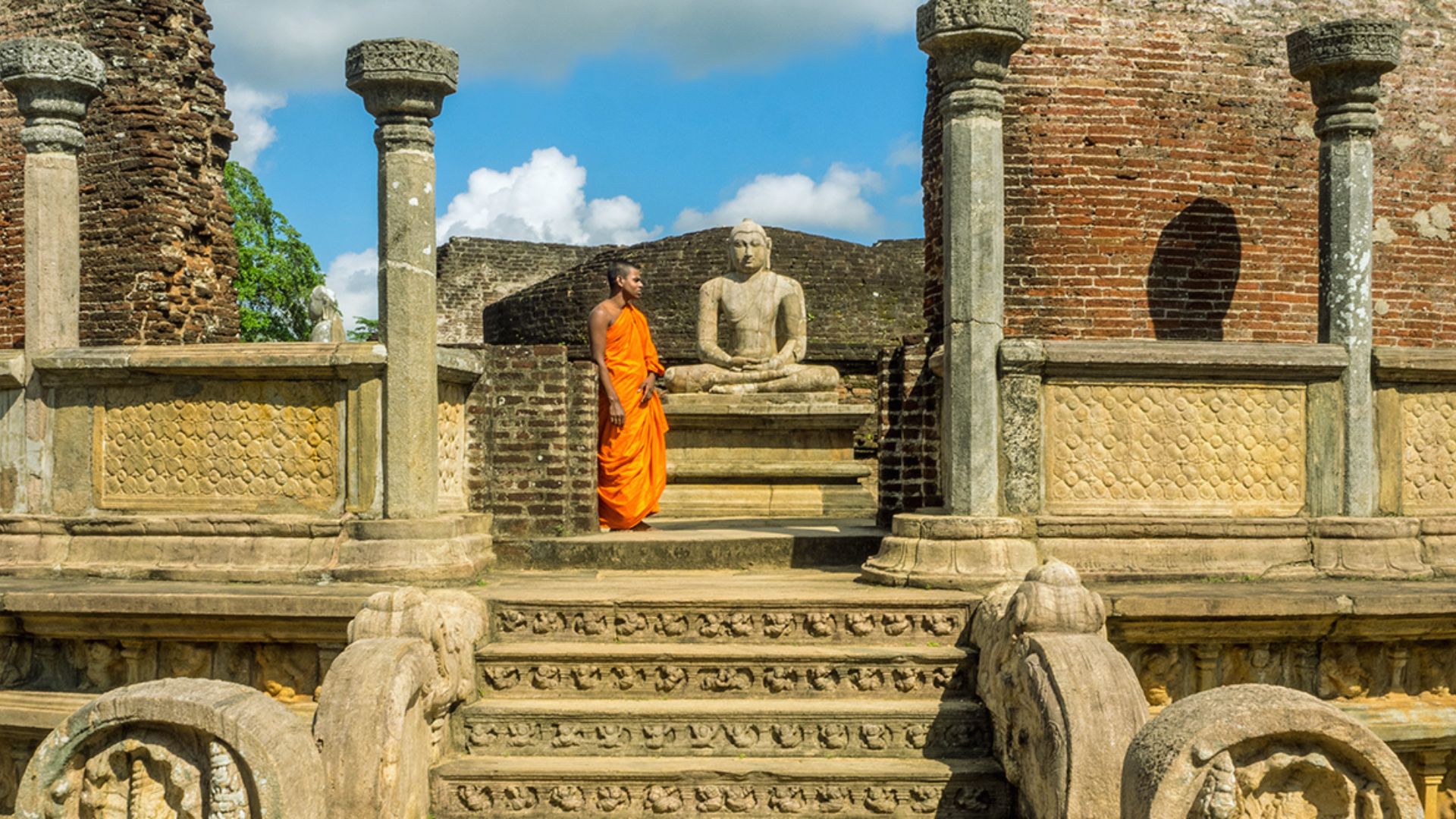 A monk in an orange robe stands on the steps of an ancient temple, surrounded by intricate stone carvings.