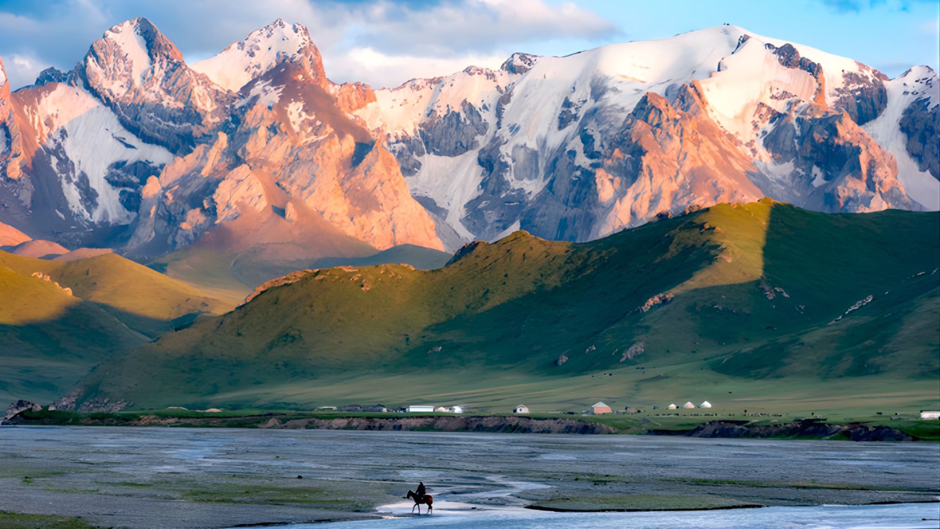 A man walks along a riverbank with mountains rising majestically in the background.