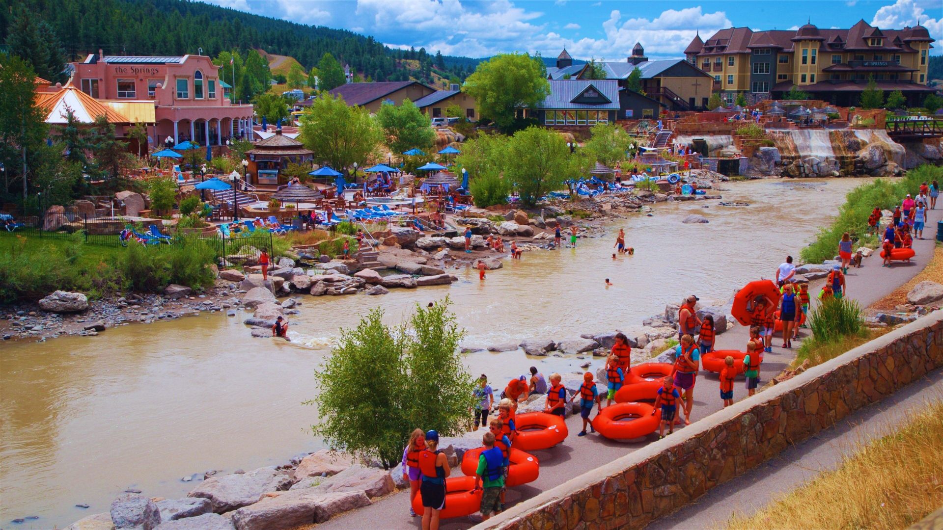 People stroll along the riverbank, passing by bright orange rafts floating on the water.