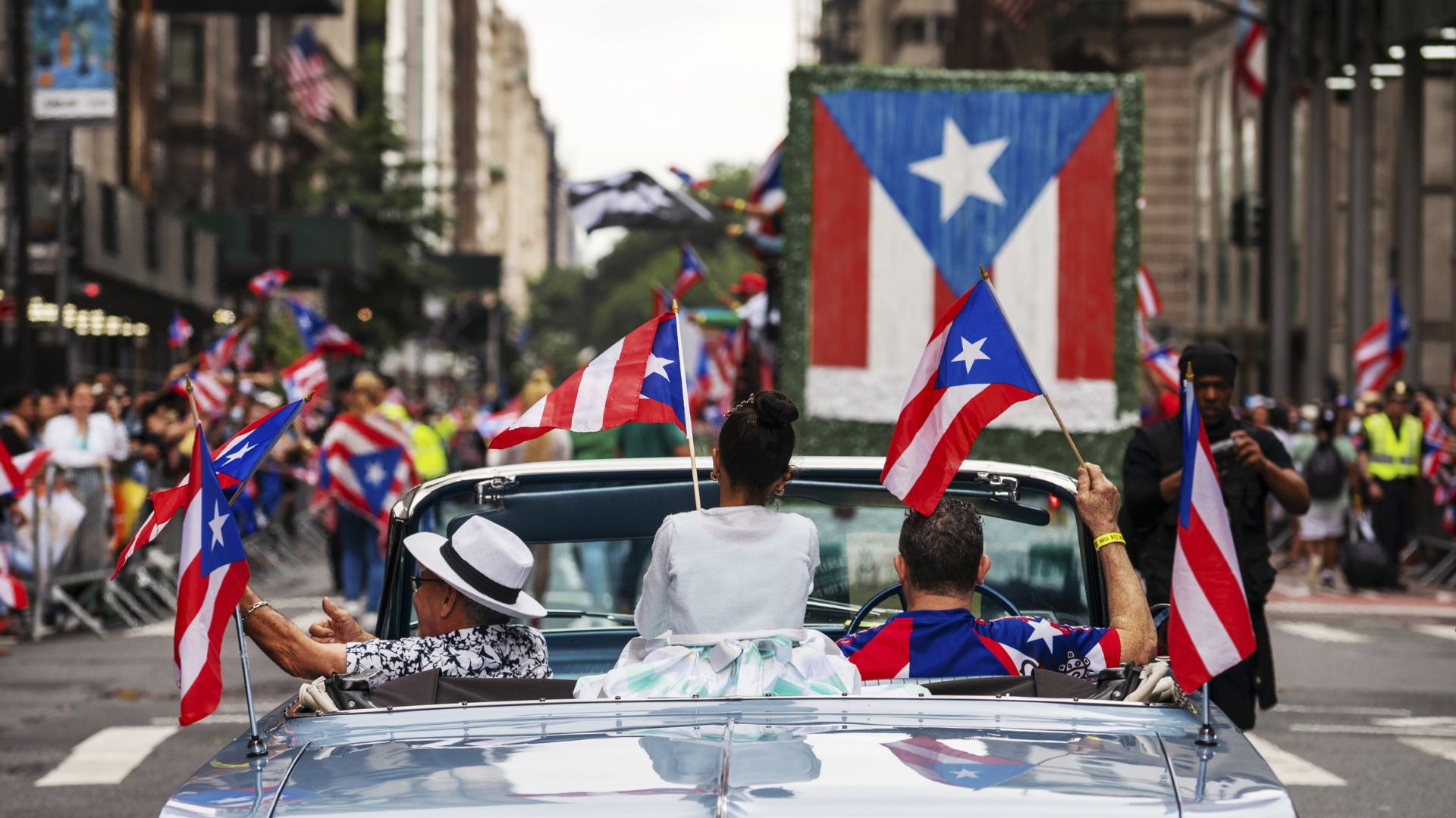 A group of individuals in a convertible car waves flags as they pass through a lively city street.