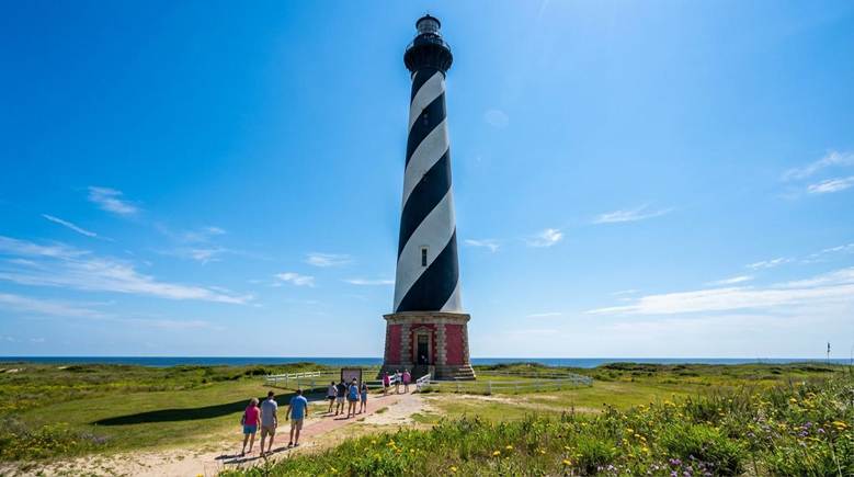 People stroll around a lighthouse on a sunny day, enjoying the bright weather and scenic views.