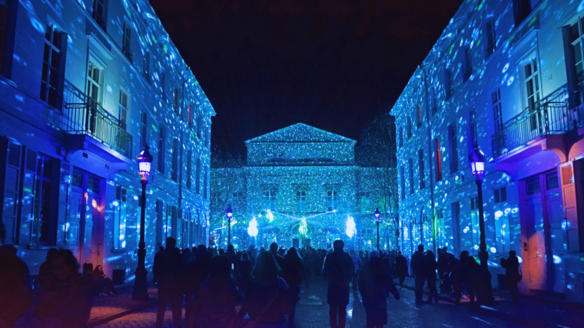 People stroll down a street illuminated by blue lights projected onto the buildings.