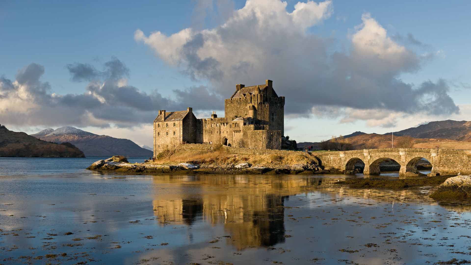 Stone castle on a small island, surrounded by a tranquil lake. The structure is connected to the mainland by an arched bridge, with mountains and a cloudy sky in the background.