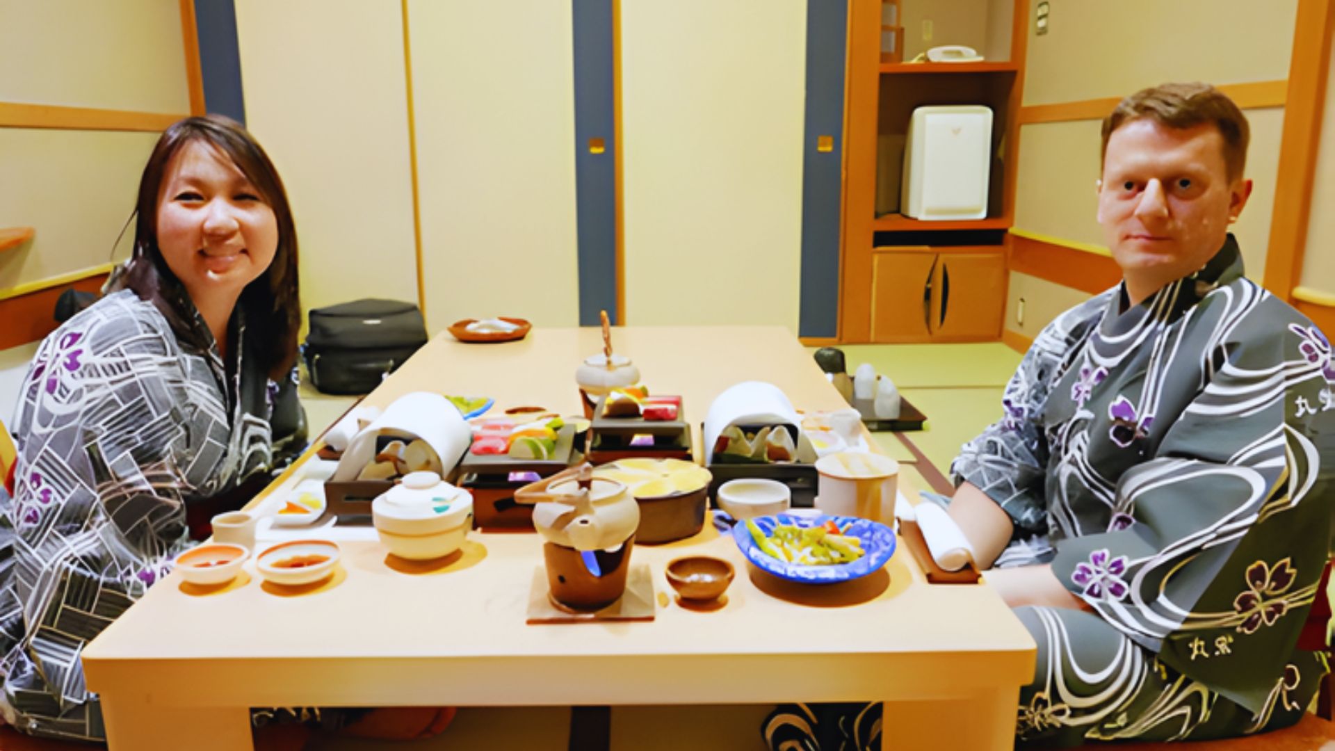 Two people in traditional robes sit at a low table with an elaborate Japanese meal in a cozy room. The setting is warm and inviting, with soft lighting.