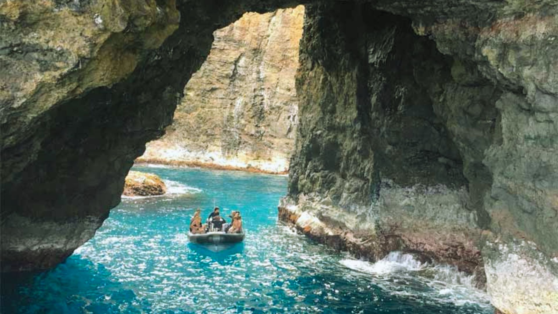 A small boat with people navigates through clear turquoise waters surrounded by towering, rugged rock formations, creating a serene and adventurous atmosphere.