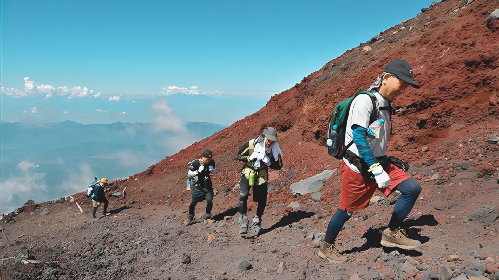 Four hikers ascend a rugged, red rocky mountain with backpacks under a clear blue sky. The scene conveys determination and adventure.