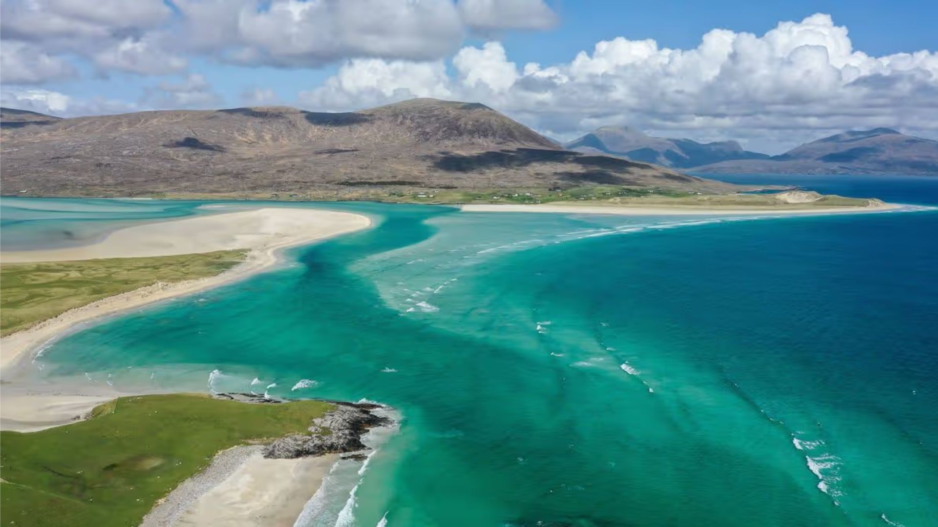 Aerial view of a turquoise coastline with sandy beaches and green fields, under a sky filled with fluffy clouds and distant mountains, conveying serenity.