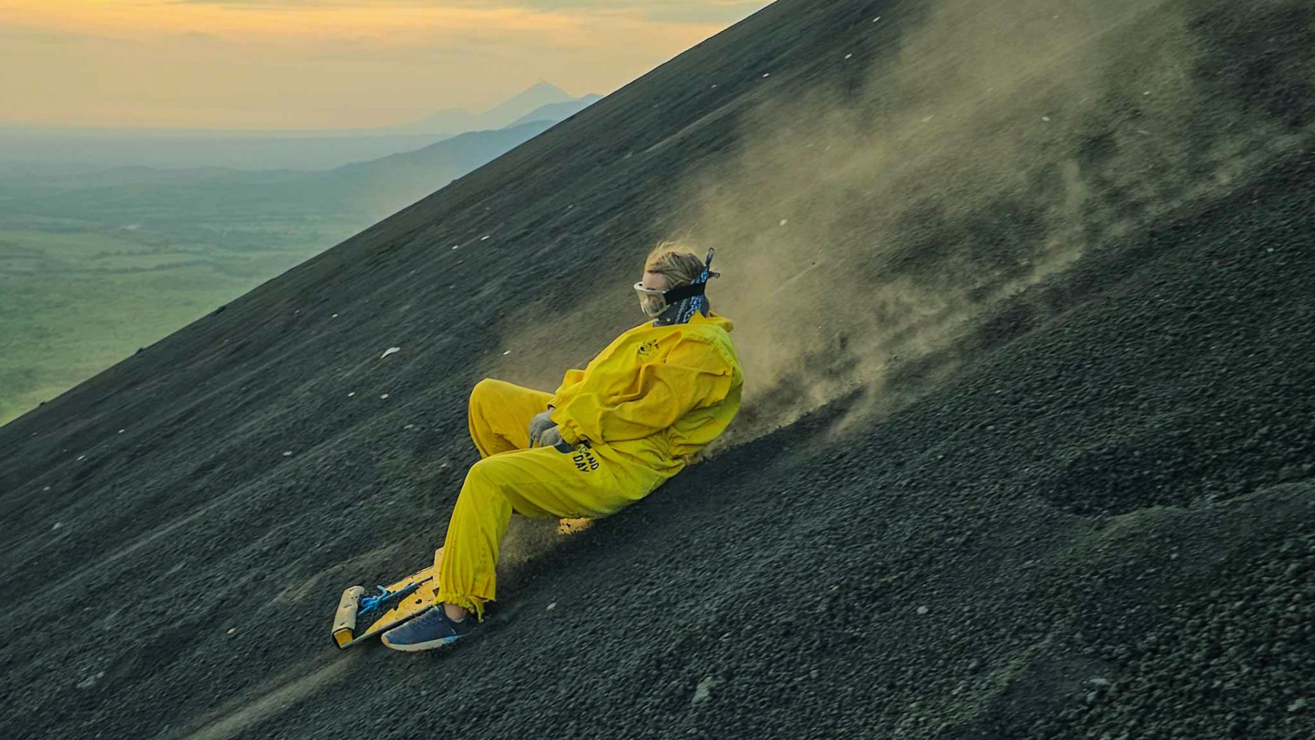 A person wearing a yellow suit and goggles slides down a steep, black volcanic slope, sending dust into the air, with a scenic view at sunset in the background.