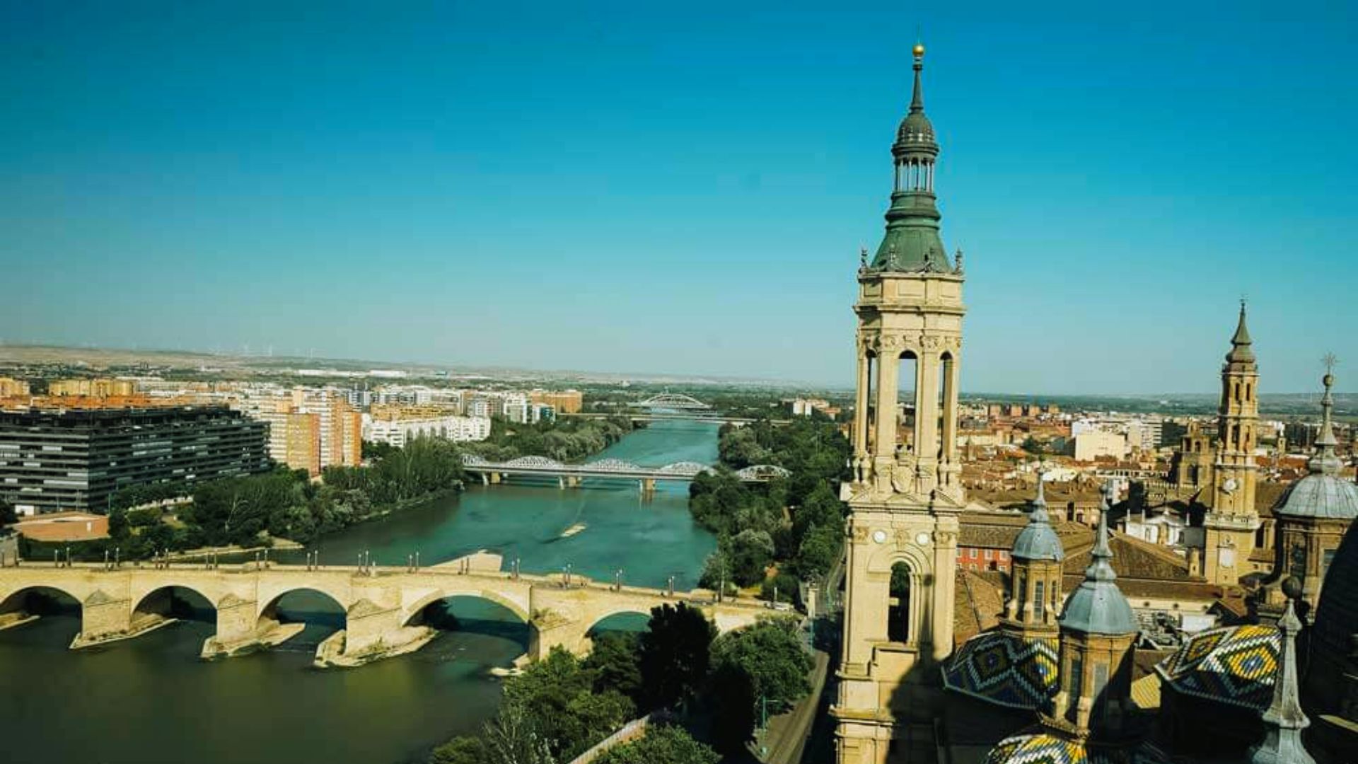 Aerial view of Zaragoza, Spain showcasing the Ebro River, stone bridges, and Basilica of Our Lady of the Pillar with its ornate towers under a clear blue sky.