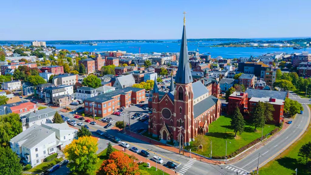 15 Beautiful Places to Visit in Maine You Can’t Miss Aerial view of a coastal town featuring a prominent red brick church with a tall steeple. Surrounding are colorful houses, trees, and a blue harbor.