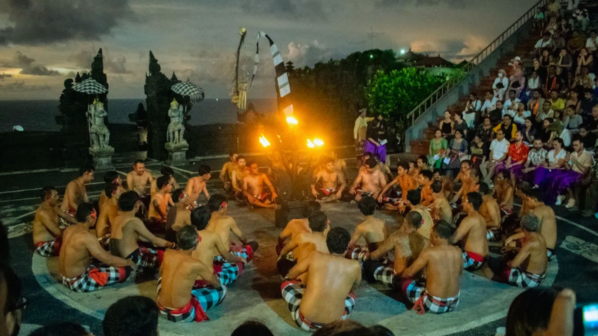 A group of people in traditional attire sit in a circle around a central fire, performing a dance at dusk, with an audience seated on the side.