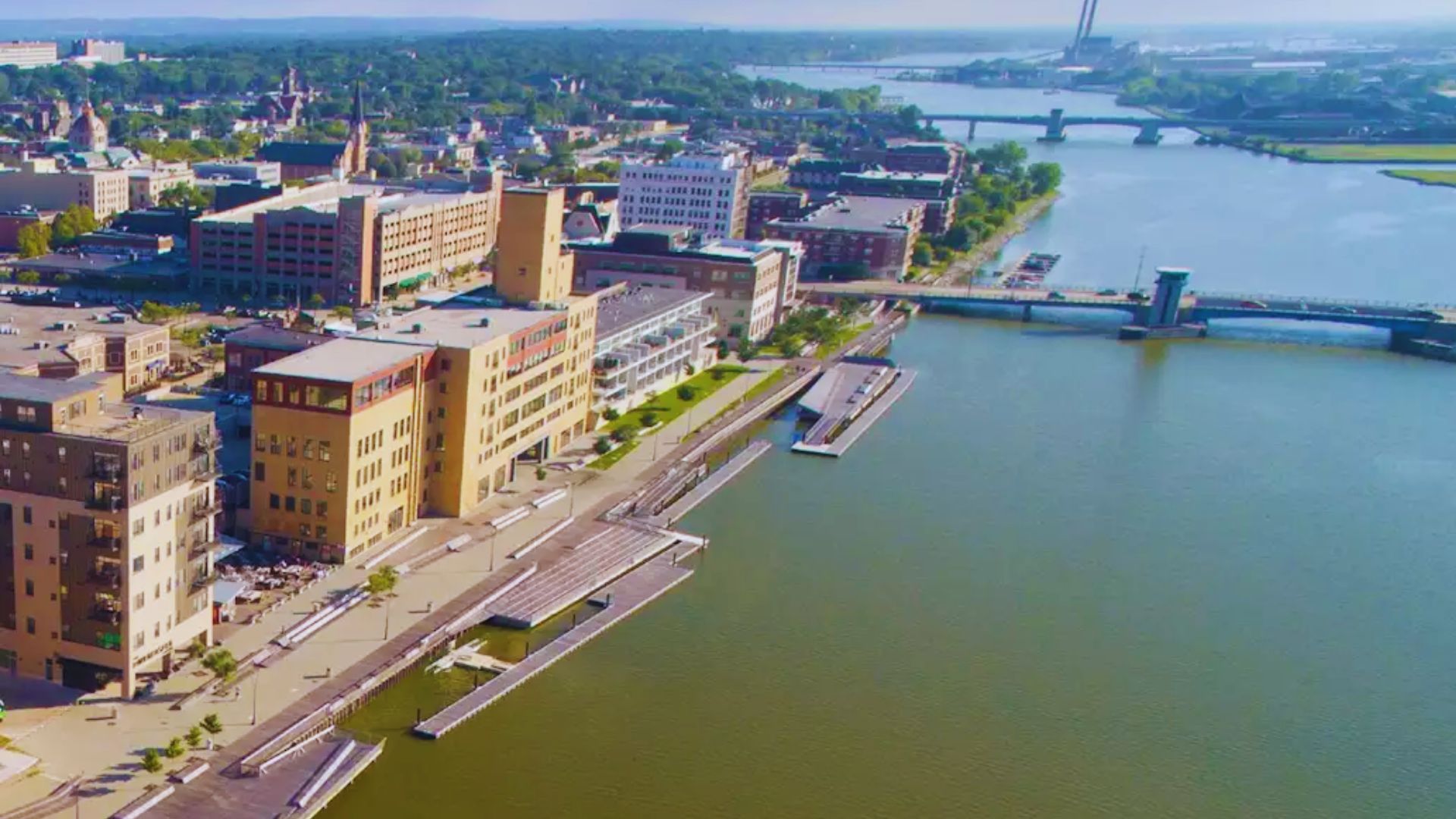 Aerial view of a waterside cityscape, featuring riverside buildings, walkways, and a bridge over a calm river, under a clear blue sky.