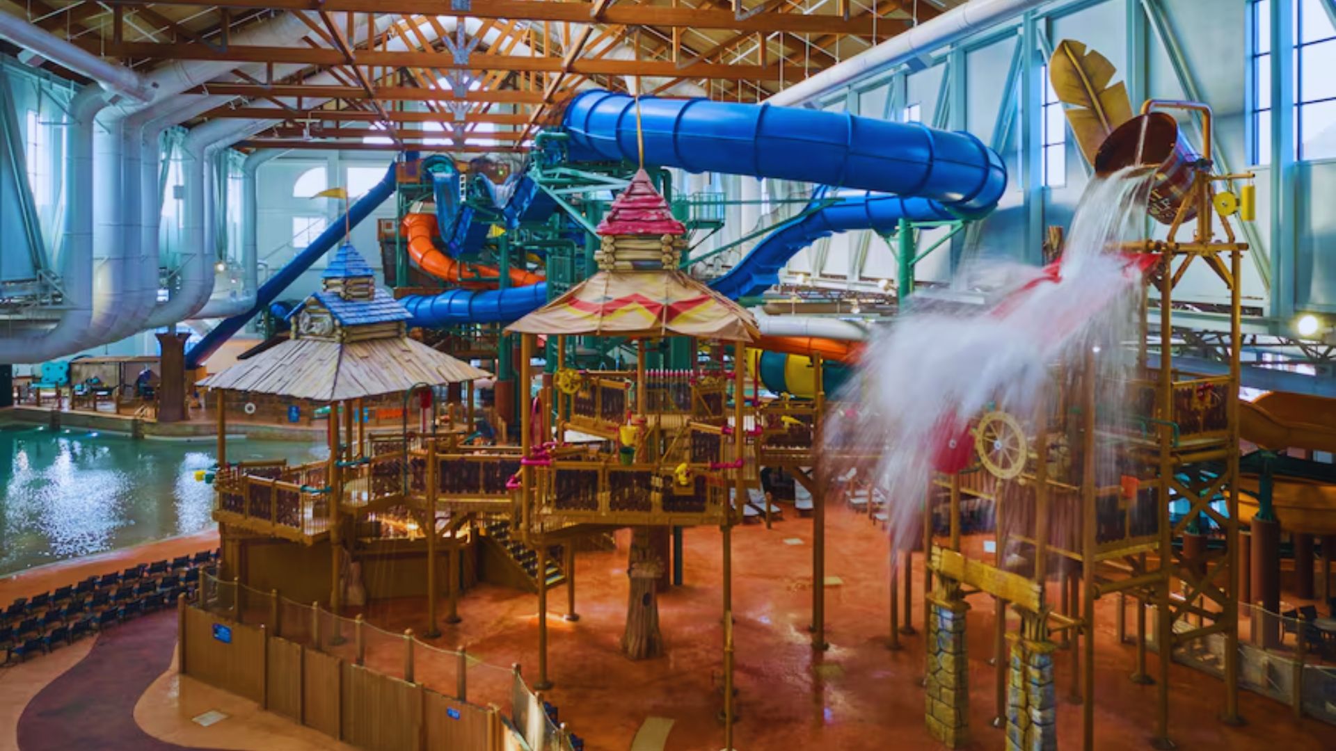 Indoor water park with twisting blue and orange slides, a large water play structure with wooden beams and colorful roofs, and water splashing from a giant bucket.