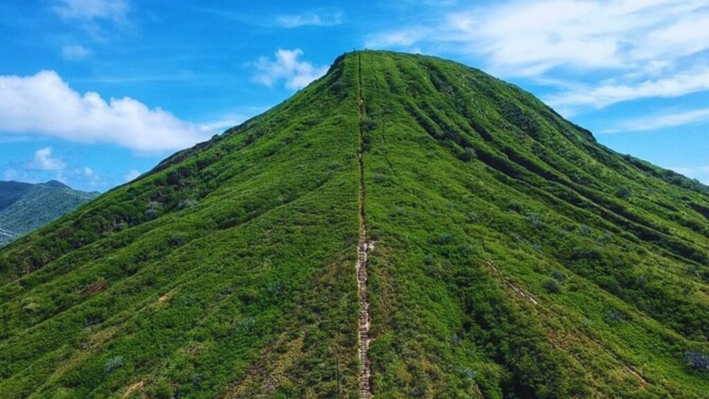 A lush, green mountain under a bright blue sky with fluffy clouds. A narrow trail runs straight up the center, creating a sense of challenge and adventure.