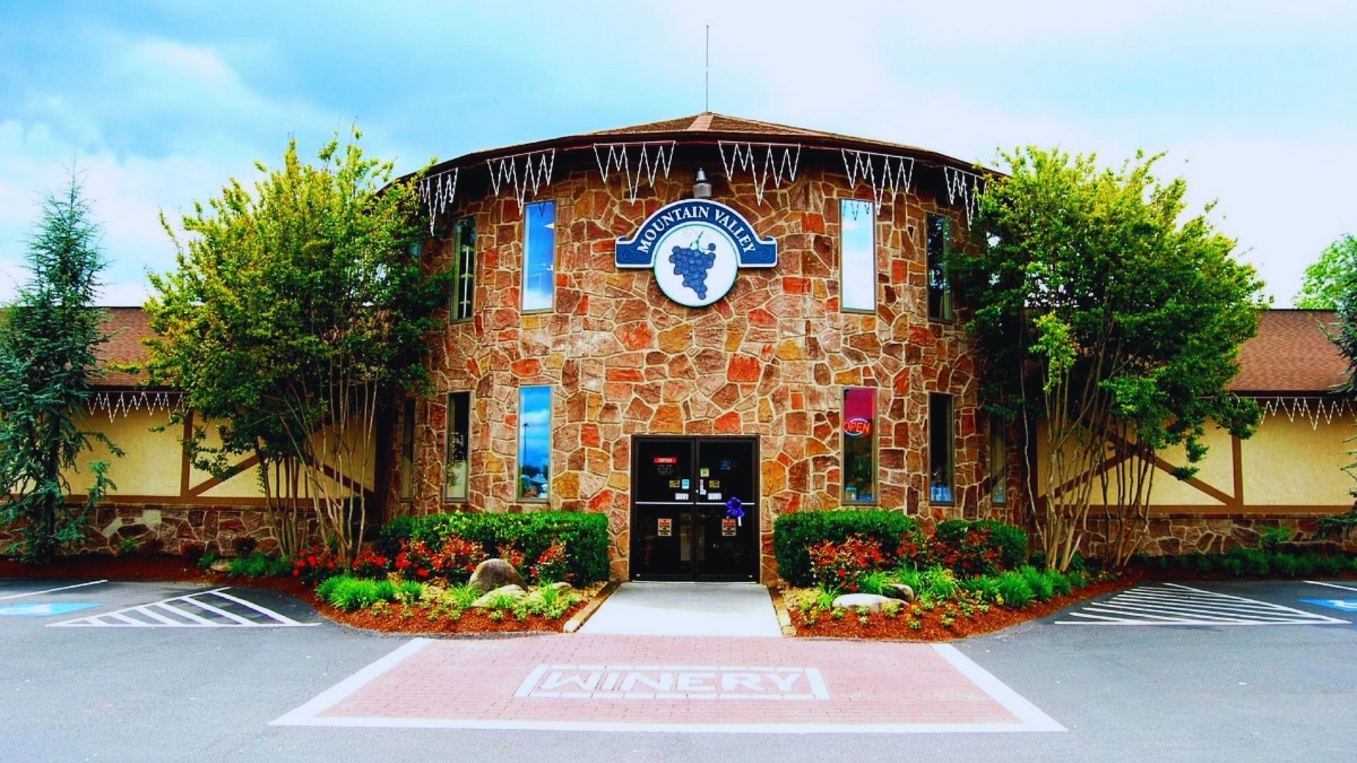 Rustic stone winery with lush greenery and a cloudy sky. Central entrance marked by a circular sign with grapes, evokes a welcoming, serene ambiance.
