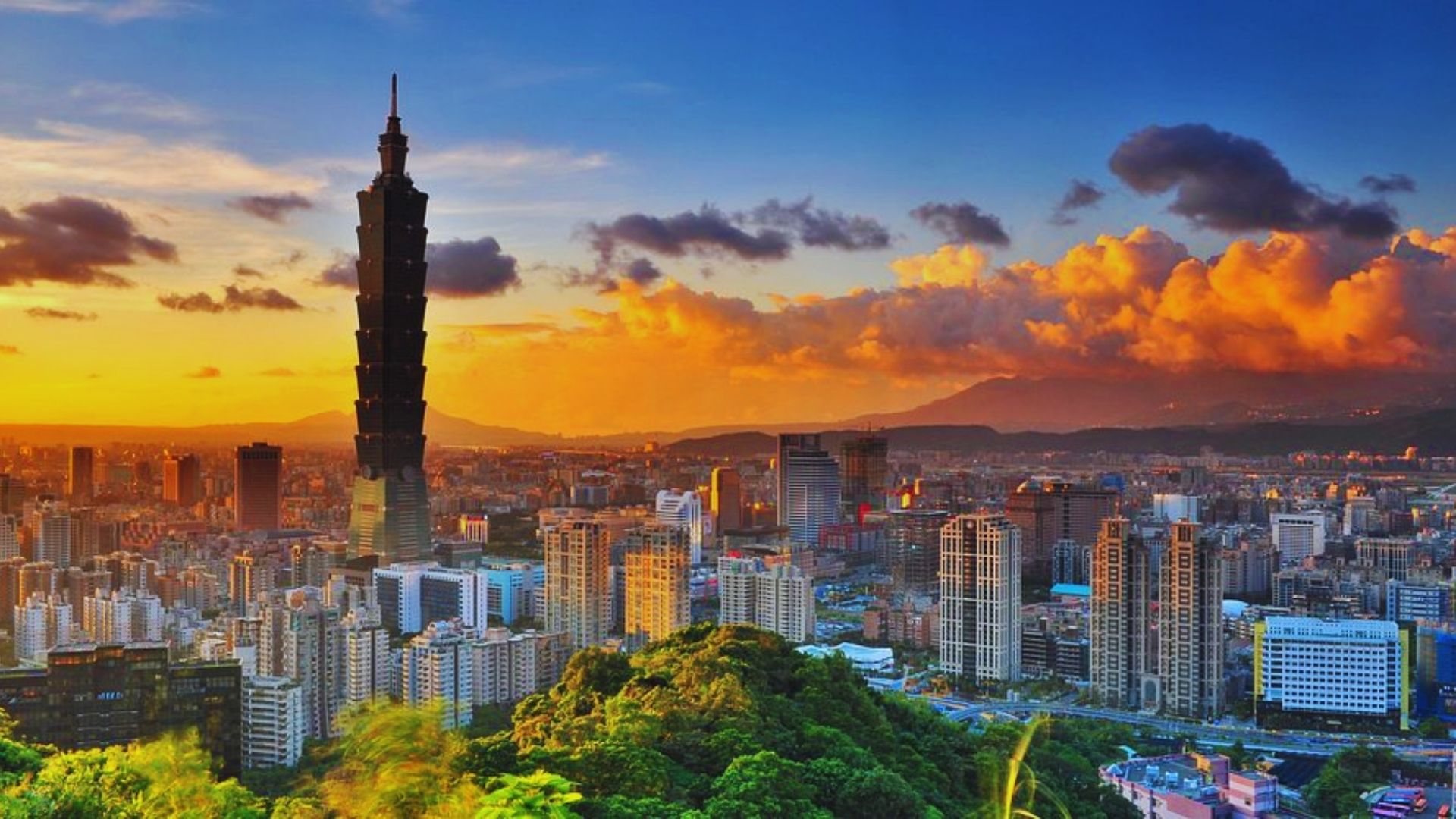 A vibrant skyline of Taipei at sunset, featuring the towering Taipei 101 skyscraper. Lush green hills in the foreground under a dramatic orange sky.