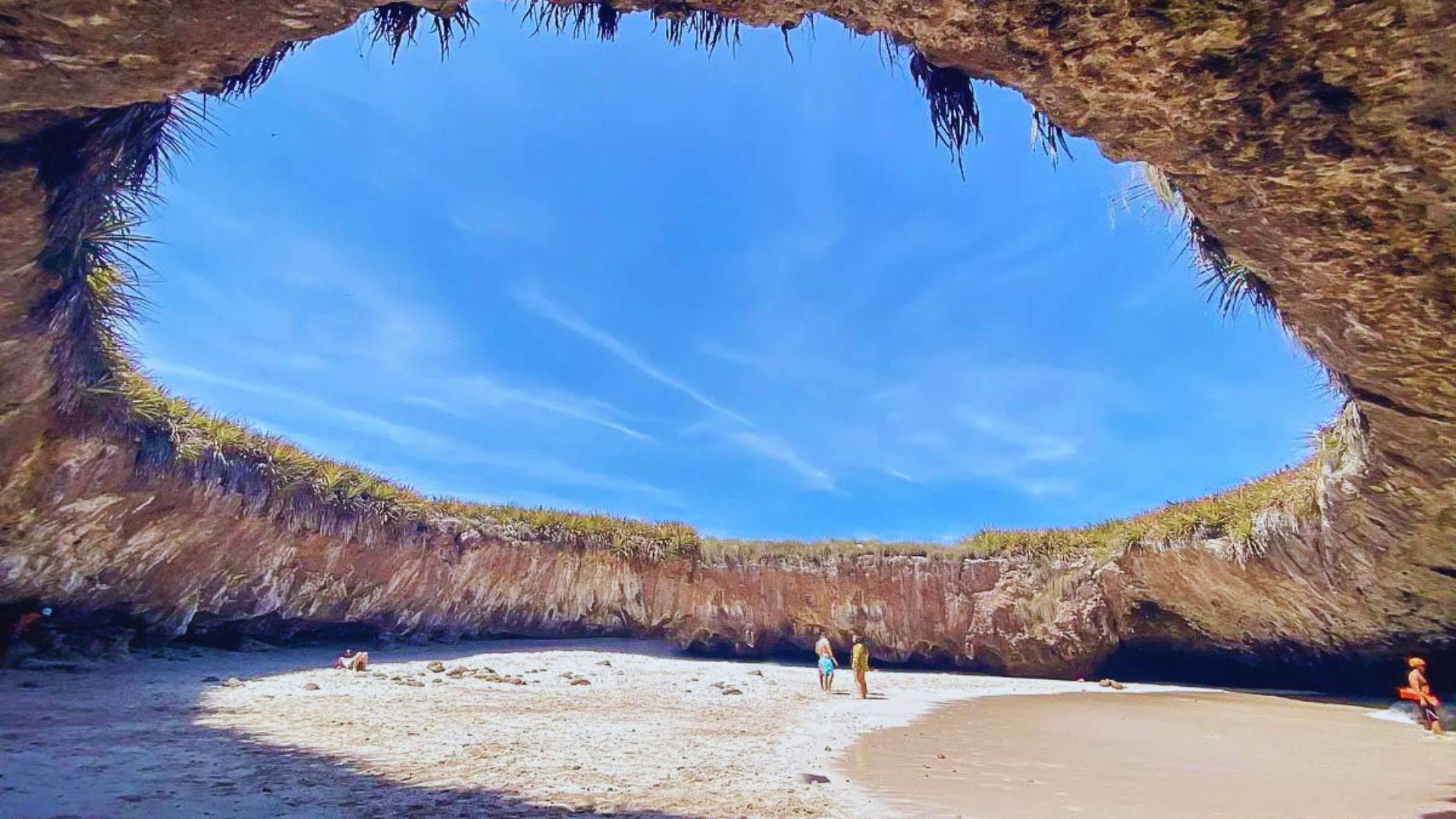 A secluded beach with a natural stone ceiling, creating an open-roof cave. Blue sky visible above; people explore the sandy area, conveying tranquility.