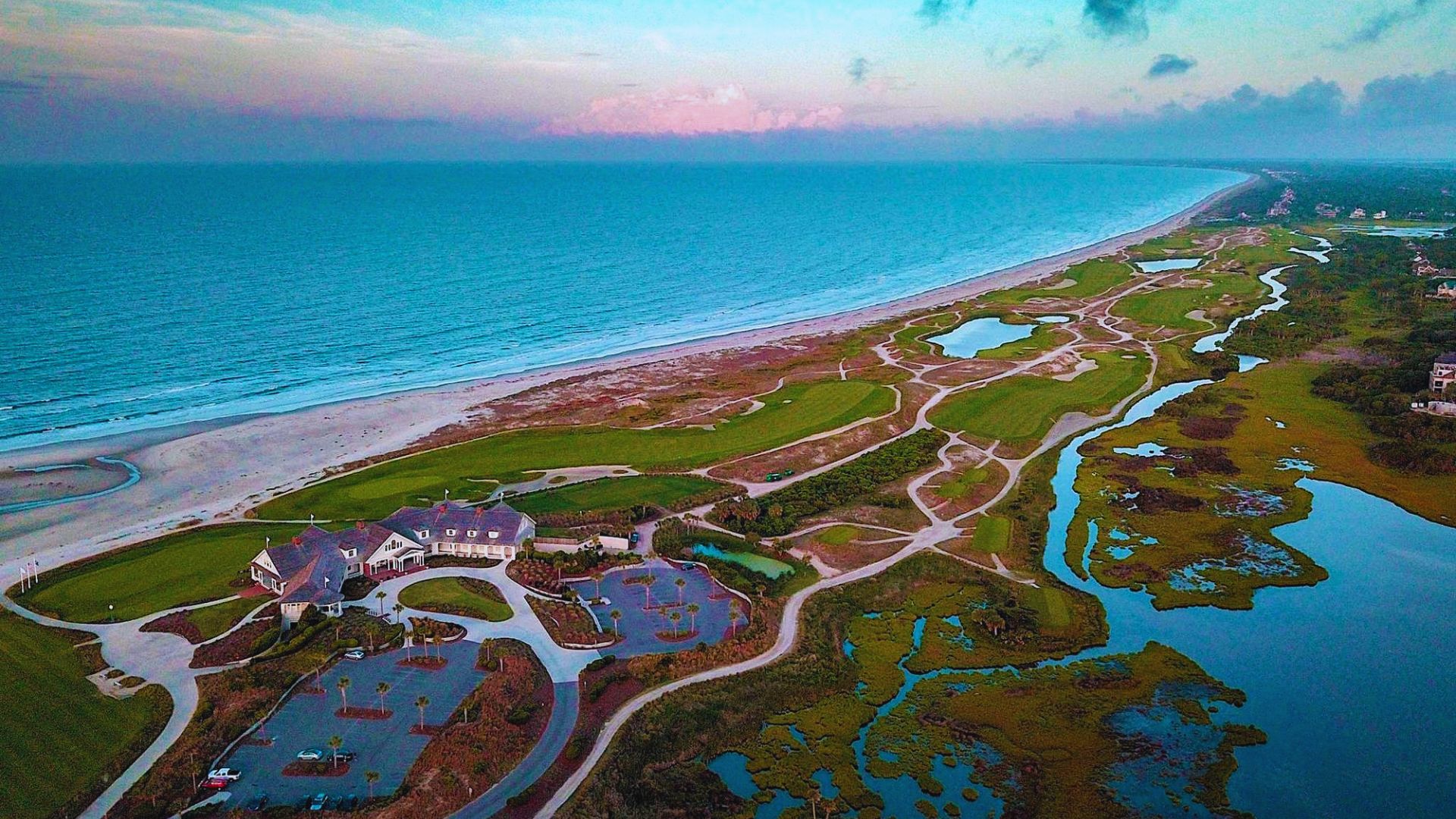 Aerial view of a coastal golf course at sunset with a sprawling clubhouse, lush green fairways, sand dunes, a calm ocean, and a pastel sky.