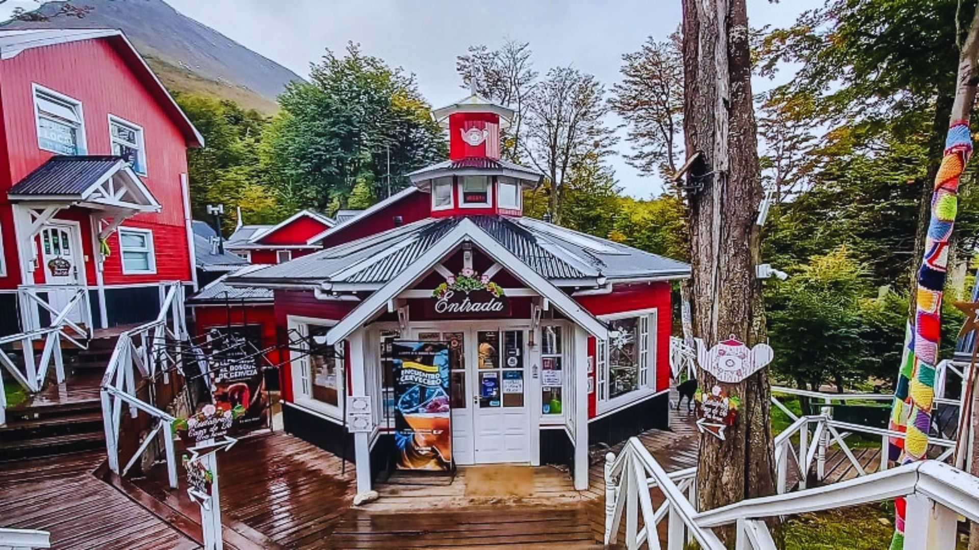 Colorful café with red walls and white trim, nestled in a forested area. Wooden walkways surround the cozy building, evoking a welcoming, rustic charm.