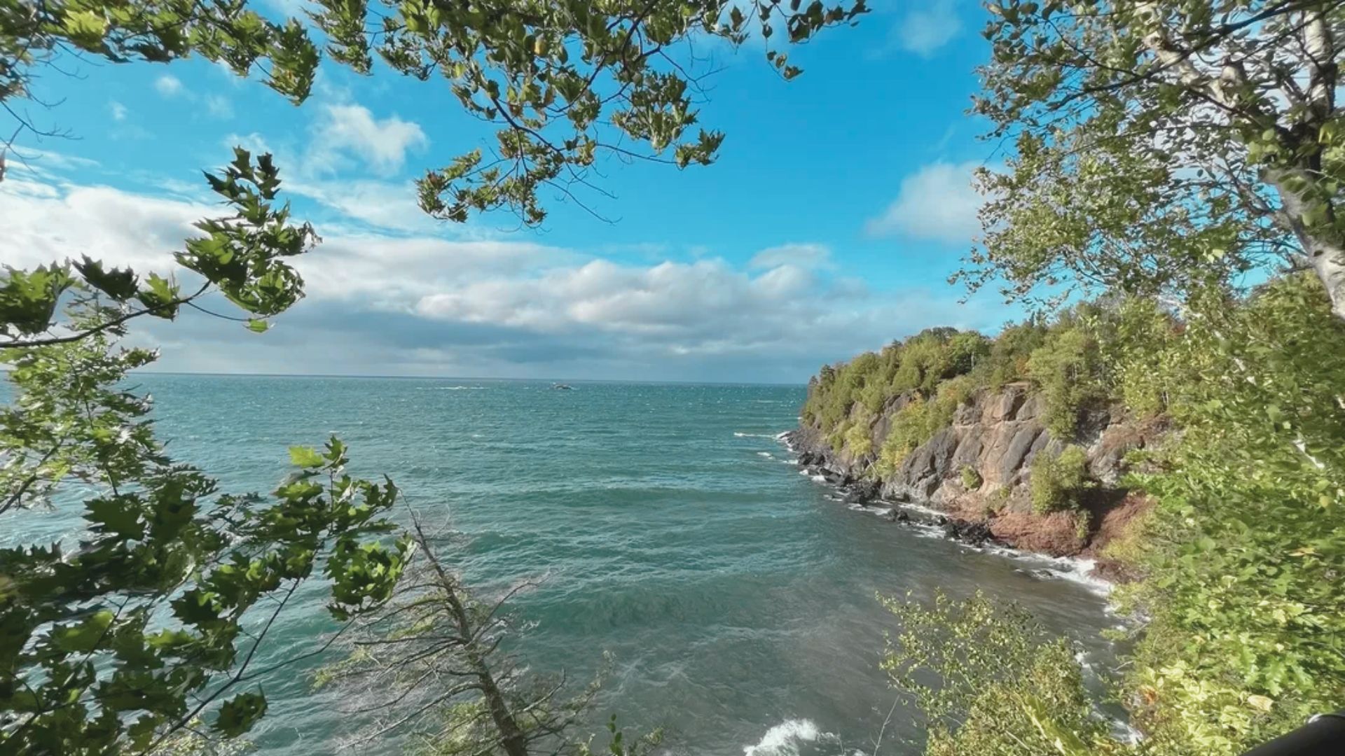 A scenic view of a rocky coastline with lush green trees framing the image. The ocean waves gently hit the shore under a bright blue sky with fluffy clouds.