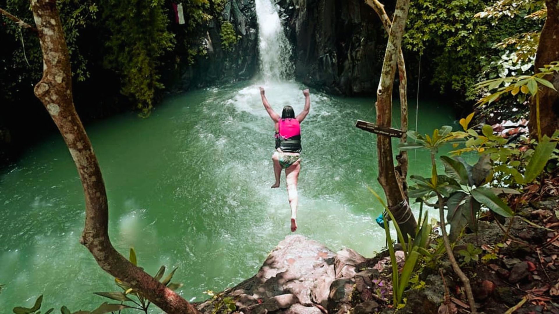 A person wearing a pink life jacket jumps into a clear green pond below a waterfall. Surrounded by lush greenery, the scene conveys adventure and excitement.