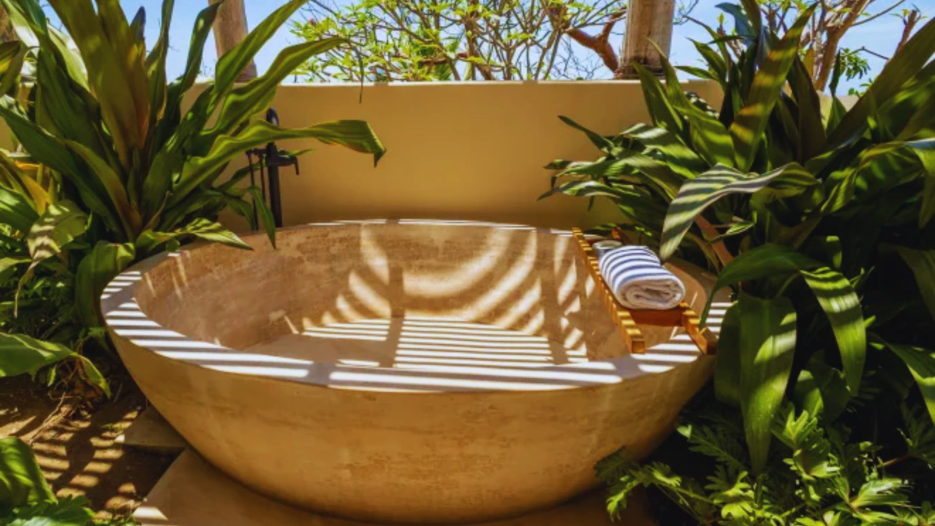 Outdoor stone bathtub surrounded by lush green foliage, with sunlight casting shadows. A wooden tray holds rolled towels, creating a tropical, serene ambiance.