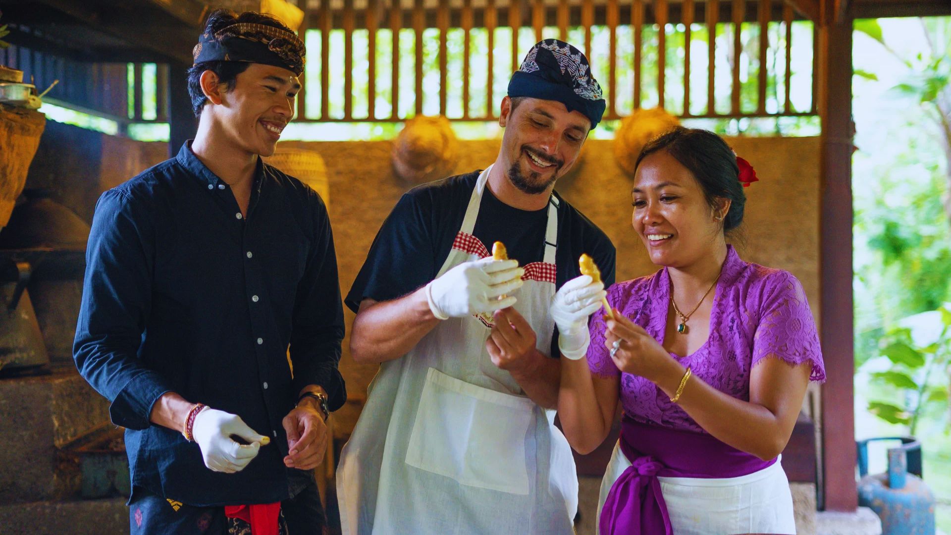 Three people in traditional attire joyfully hold food in a rustic kitchen. They wear gloves, displaying a sense of camaraderie and cultural celebration.