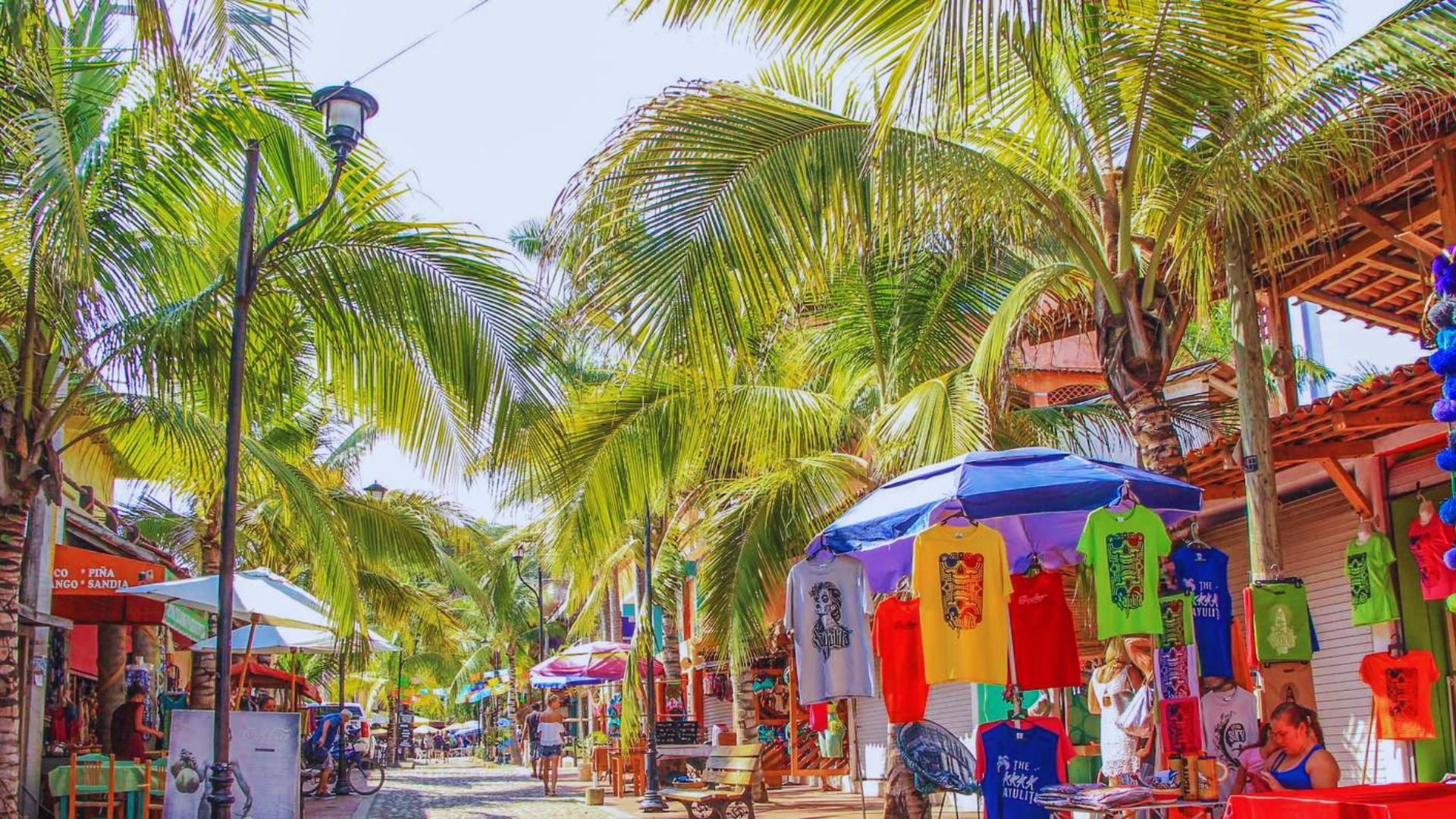 A vibrant street scene under sunny skies featuring tall palm trees casting shadows over colorful market stalls. Brightly colored T-shirts hang on display, creating a lively and tropical atmosphere.