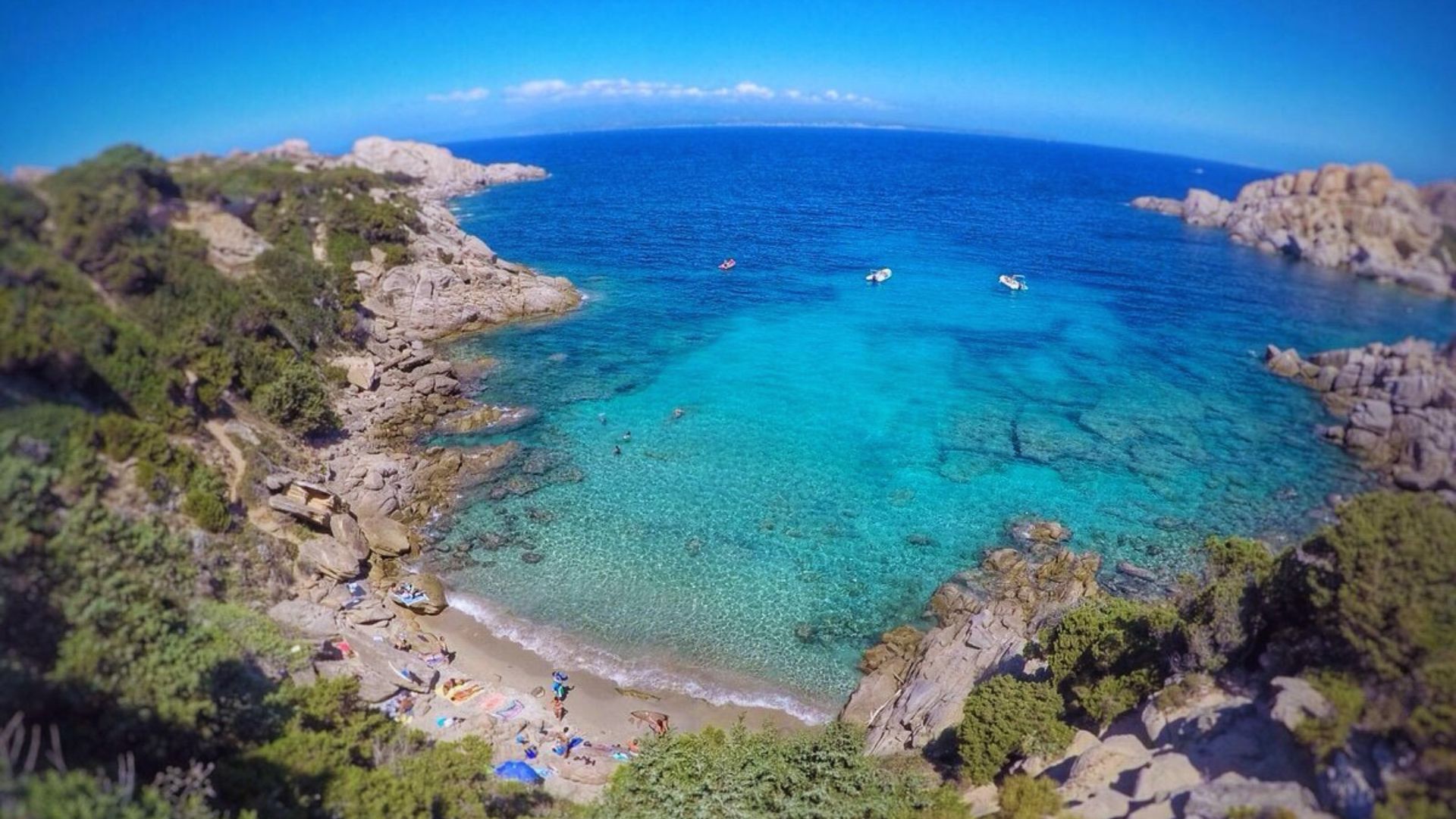 Aerial view of a serene cove with turquoise waters, rocky cliffs, and lush greenery. People relax on the sandy beach, and boats float nearby.