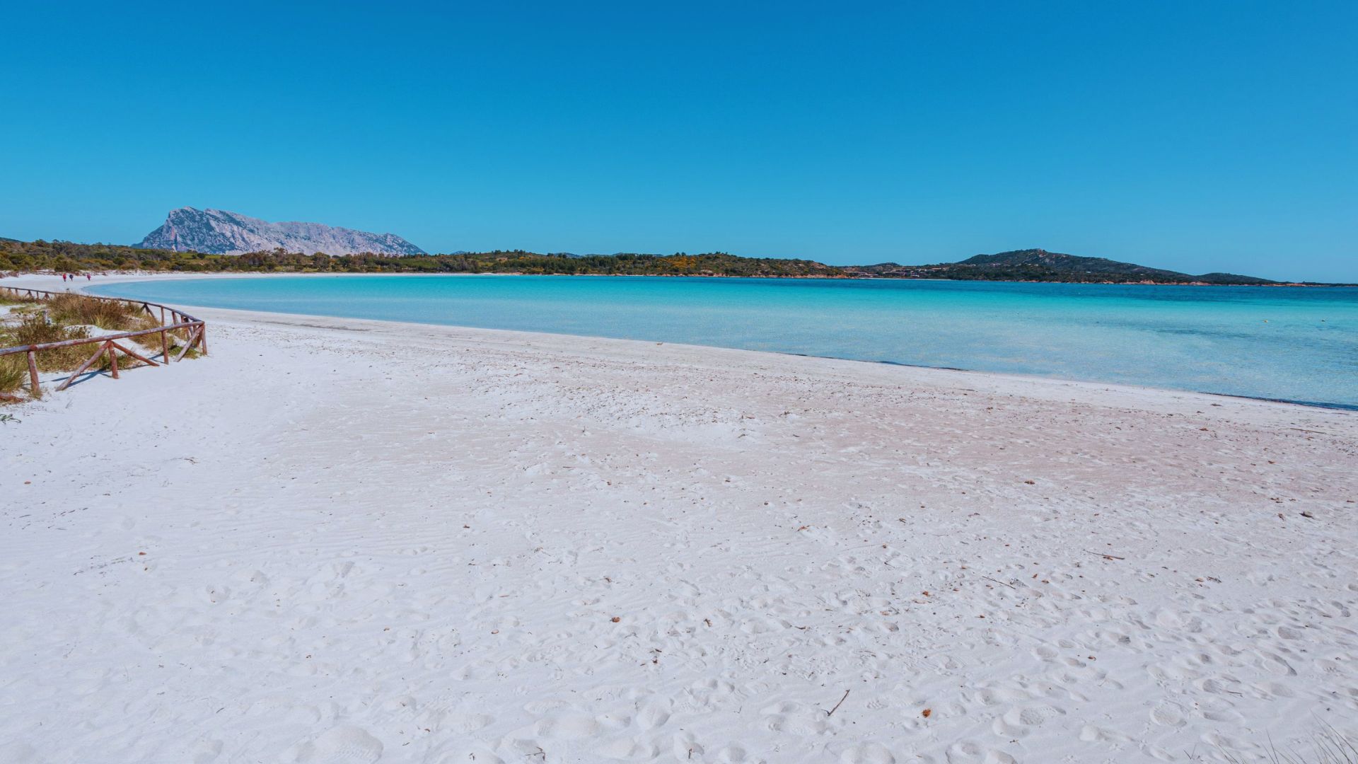 Serene beach scene with white sand, turquoise water, and clear blue sky. Distant hills frame the horizon, evoking a sense of tranquility and openness.