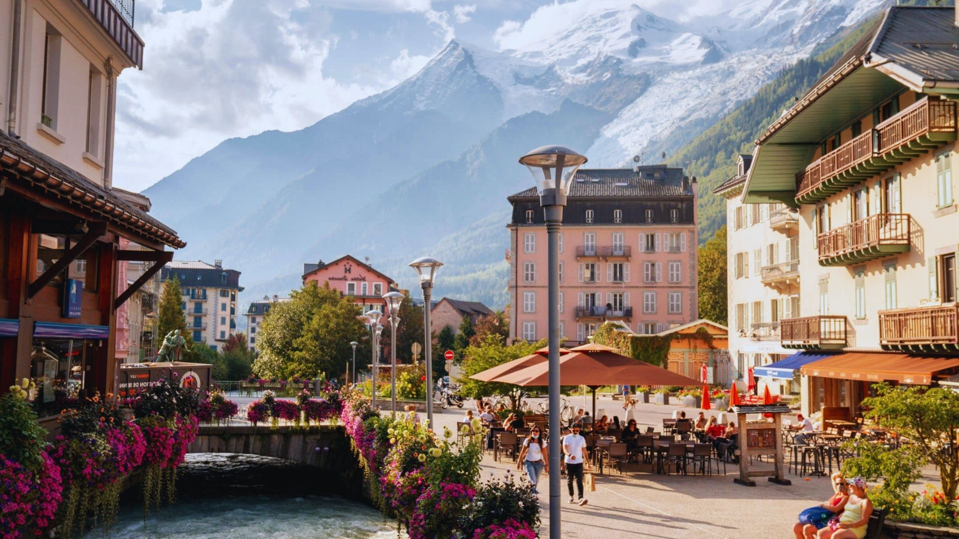 A river flows through a town, with majestic mountains rising in the background under a clear blue sky.