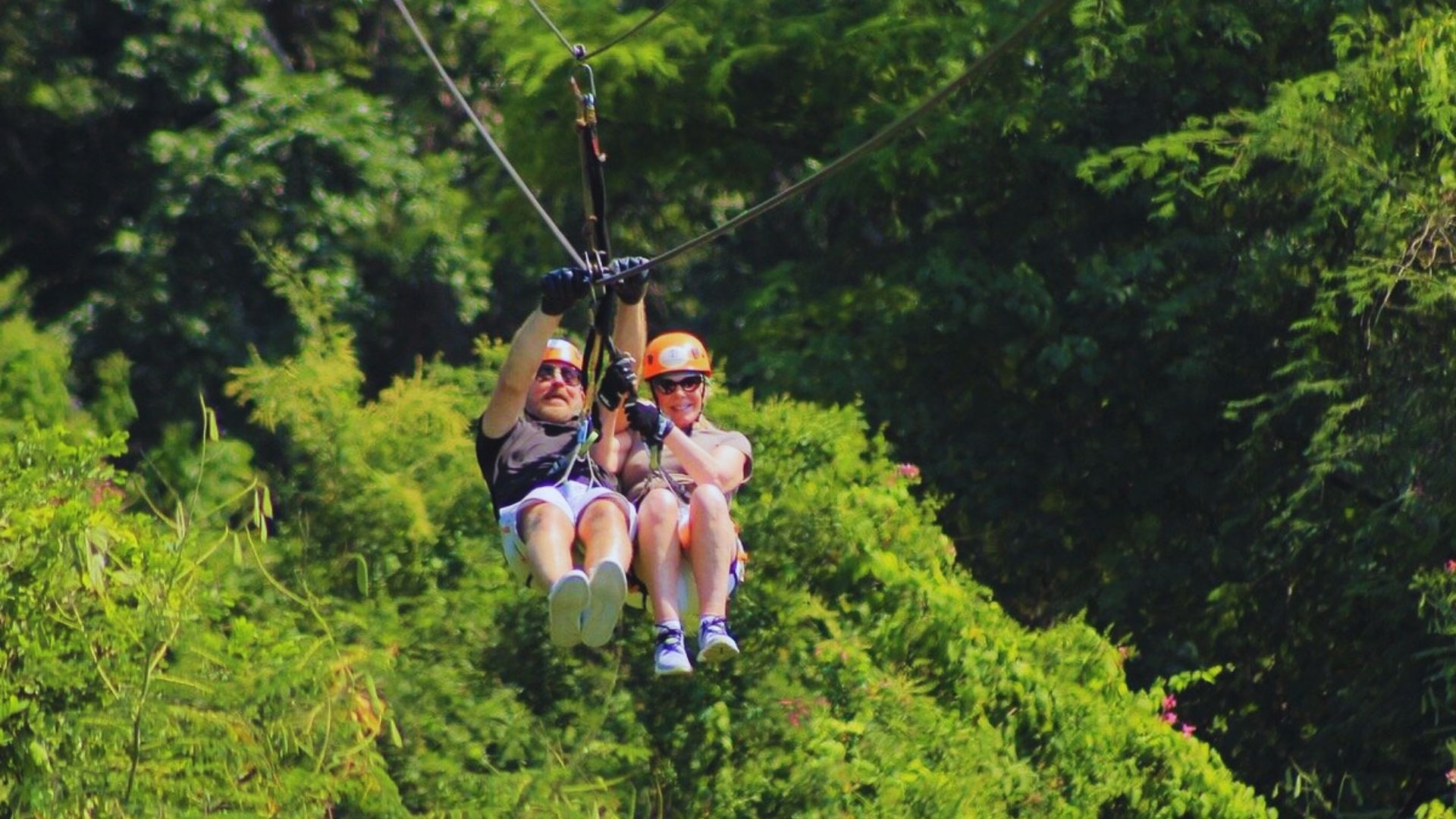 Two people zipline through lush greenery, wearing helmets and harnesses. Both show excitement, capturing a thrilling, adventurous moment amidst nature.