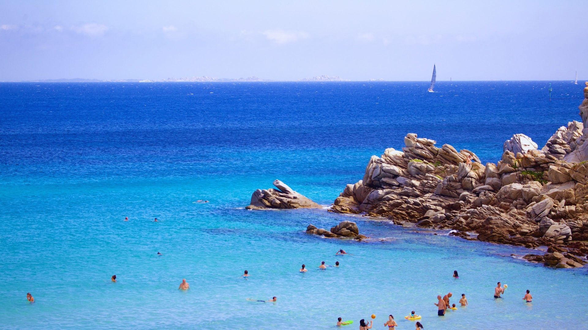 A vibrant beach scene with clear turquoise water, scattered swimmers, and rocky cliffs on the right. A sailboat glides on deep blue sea under a clear sky.