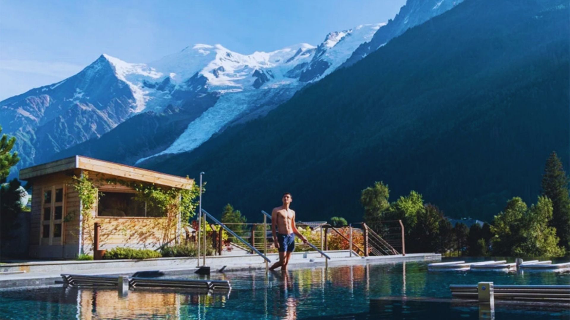 A man stands in a mountain-top pool, surrounded by breathtaking views of the peaks and sky.