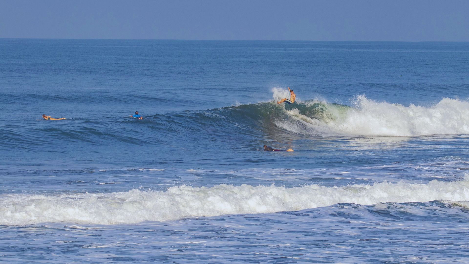 Surfer rides a small wave on a bright blue ocean, with others in the water. The scene exudes a relaxed, adventurous vibe under a clear sky.